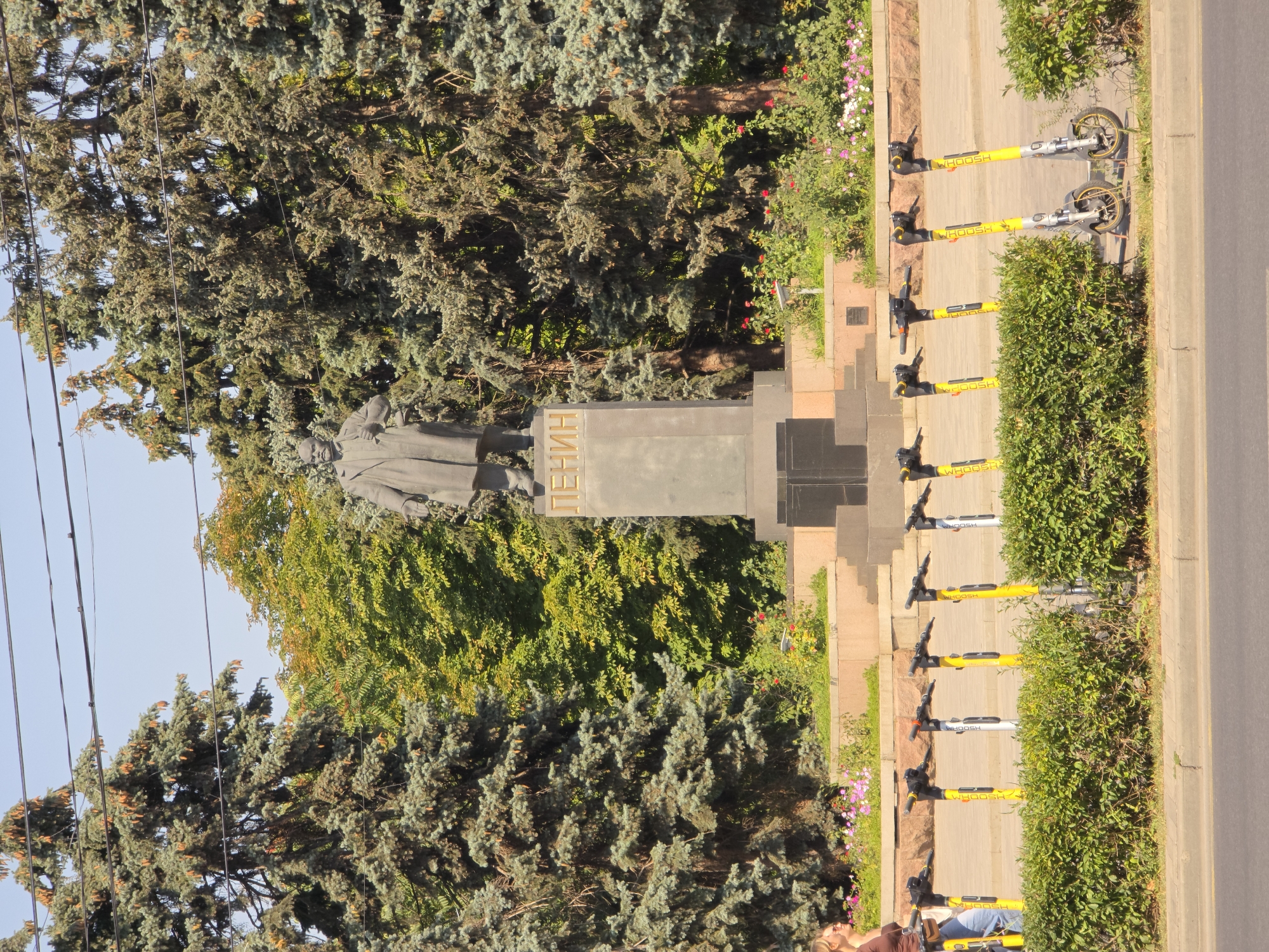 The image depicts a statue of Vladimir Lenin situated in a park in Rostov-on-Don, Russia. The statue, made of bronze or stone, stands on a tall granite pedestal. The pedestal is inscribed with the name "ЛЕНИН" (Lenin) in golden Cyrillic letters. The statue is flanked by tall evergreen trees, with a deciduous tree displaying some autumn foliage behind it. In front of the pedestal, a low, manicured hedge borders a walkway. Beyond the hedge, a row of yellow electric scooters is parked, several of which bear the "WHOOSH" logo. A woman, dressed in a brown top and blue jeans, walks past the scooters. In the background, a paved area with flowerbeds containing pink and white flowers adds to the park-like setting. The sky is clear and blue, indicating a sunny day. The scene appears to be a public space, possibly a square or a park in the city.