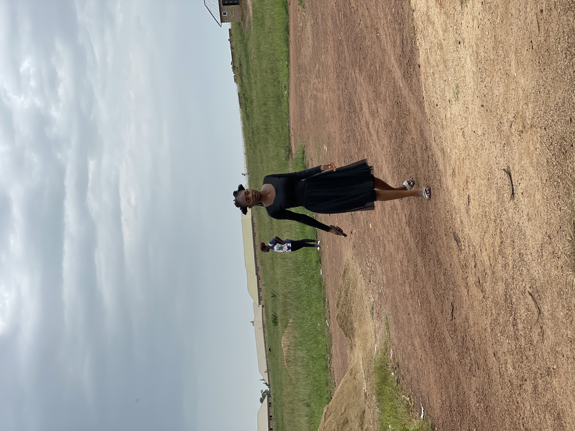 A young Black woman in a black dress walks on a dirt road in Durumi, Nigeria, under a cloudy sky. She has her hair in two buns. She is holding a dark object in her right hand, possibly a phone. To her left, a pile of sand is visible. In the background, there are buildings with yellow roofs and a grassy field. Another person in a t-shirt and leggings stands in the grassy area. The scene is outdoors and appears to be a semi-rural or developing area.