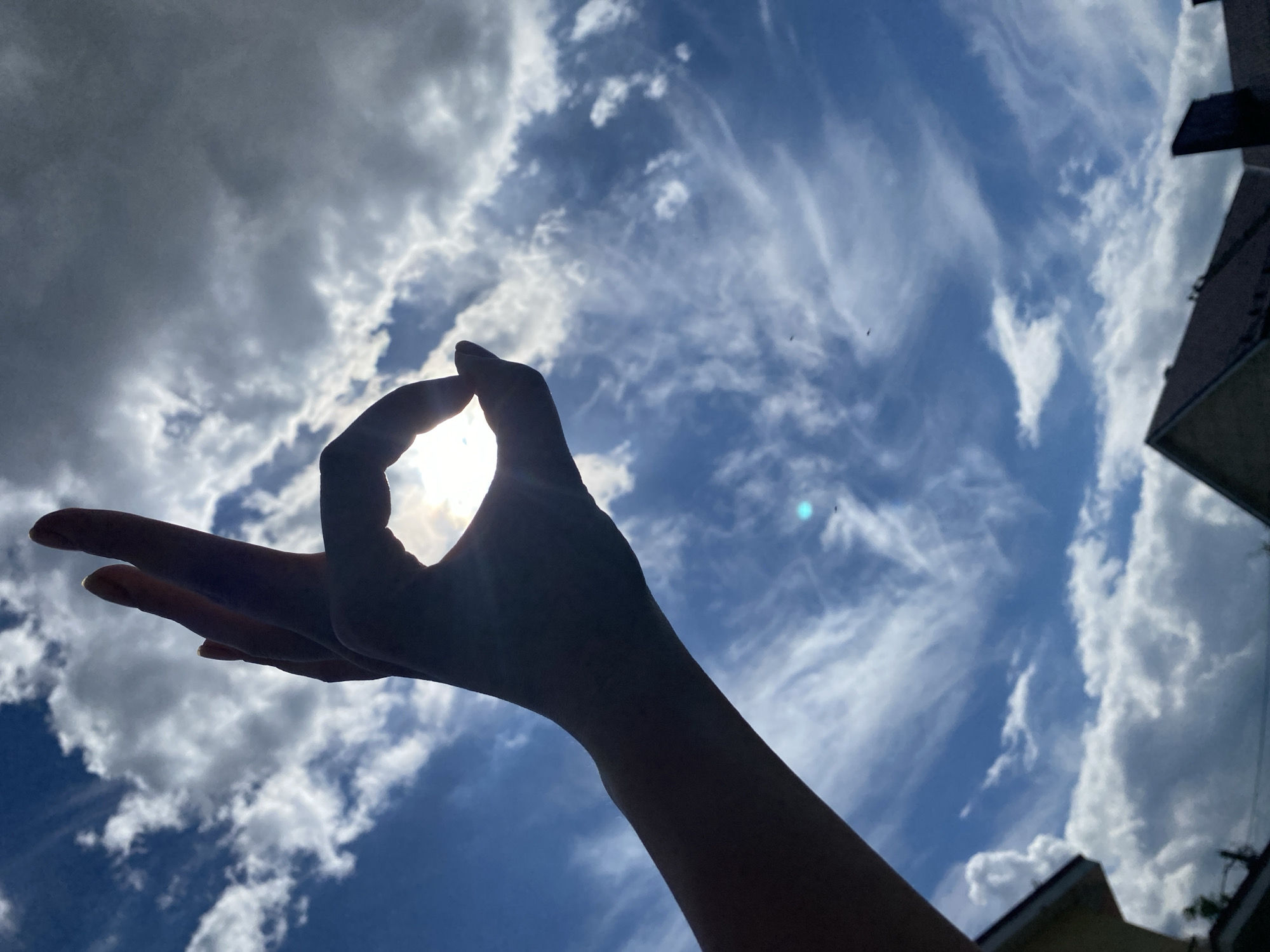 The media file displays a silhouette of a human hand against a bright sky. The hand is positioned in the foreground, with the thumb and index finger forming a circle, possibly holding or framing the sun which is visible behind it. The sun's rays are casting light through the opening, creating a halo effect. The rest of the hand is outstretched, with fingers extended.
The background is a vibrant blue sky filled with dramatic clouds. Some clouds are dense and white, suggesting cumulus formations, while others are thin and wispy, streaking across the sky like cirrus clouds. The interplay of light and shadow on the clouds indicates a dynamic sky, possibly during daytime.
In the upper right corner of the image, the dark silhouette of a building or structure is visible, providing a hint of an urban or semi-urban setting. The angle of the shot suggests it was taken from a low vantage point, looking upwards.
The overall impression is one of contemplation or mindfulness, as the hand gesture is often associated with meditation or yoga. The bright light and expansive sky evoke a sense of openness and perhaps optimism. The image captures a moment of connection between the human form and the natural elements, with the sun acting as a focal point. There is no visible text in the image.