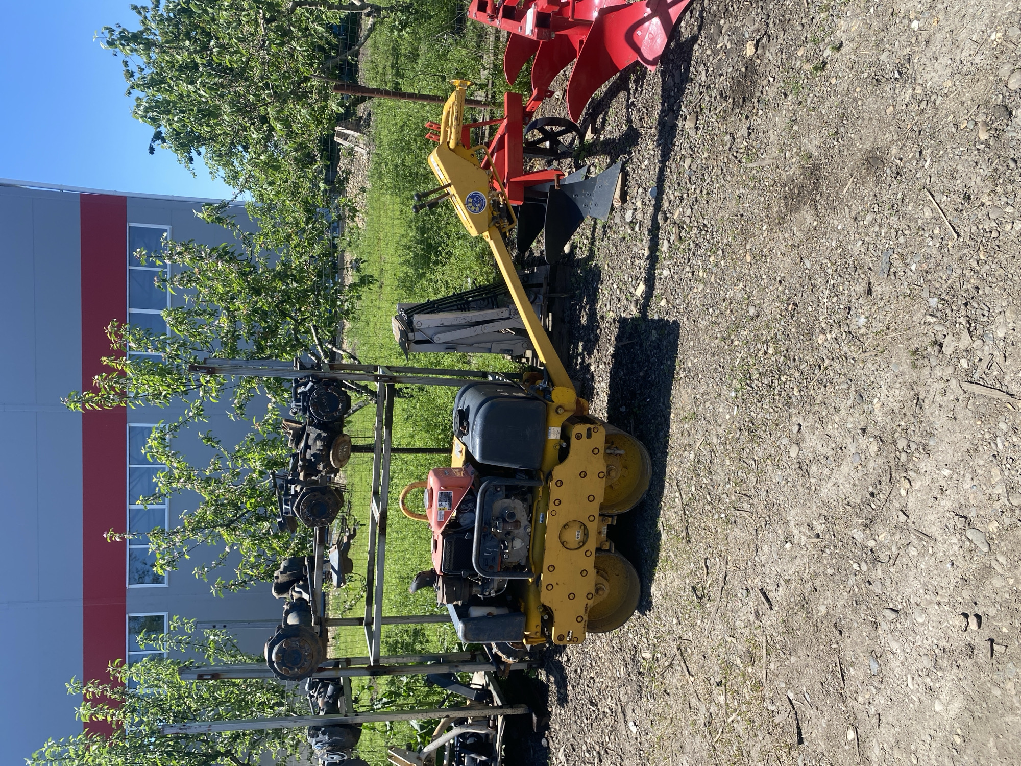 The image shows an outdoor scene in what appears to be a storage area or a yard adjacent to a building. The building is light gray with a wide red stripe. The sky is clear blue.

The primary objects of interest are agricultural and construction machinery. A yellow double-drum vibratory roller compactor is prominently displayed near the center. To its left is a metal rack supporting several pieces of machinery, possibly axles or other vehicle components. Further to the right and slightly behind the roller, a red plow attachment is visible. A yellow cultivating tool appears alongside the plow.

The ground is covered in gravel and dirt. Patches of grass and trees are scattered throughout the background.

The location is indicated to be in the city of Neftyanik, Russia. However, there are no specific visual cues or signage in the image to confirm this location definitively.
