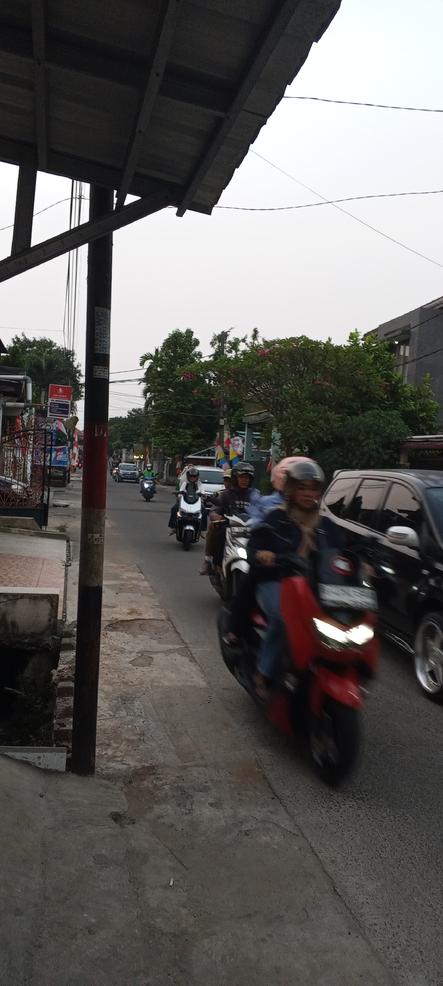 The image depicts a street scene in Depok, Indonesia, possibly during the late afternoon or early evening, given the ambient light. A busy street is visible with several motorcycles and a car. In the foreground, a person on a red motorcycle is in motion, their face partially obscured by a helmet. To their left, a black car is also moving. Further down the street, other motorcycles are visible, carrying riders also wearing helmets. The road surface appears to be asphalt with some cracks and discoloration. Along the side of the road, there is a concrete walkway and a drainage ditch. Buildings and trees line the street, with some houses and a commercial establishment identifiable. Utility poles and wires are present overhead, indicating an urban setting. Decorative flags or banners are also visible, suggesting a possible festive or communal event. The overall atmosphere suggests moderate traffic and daily life activity.