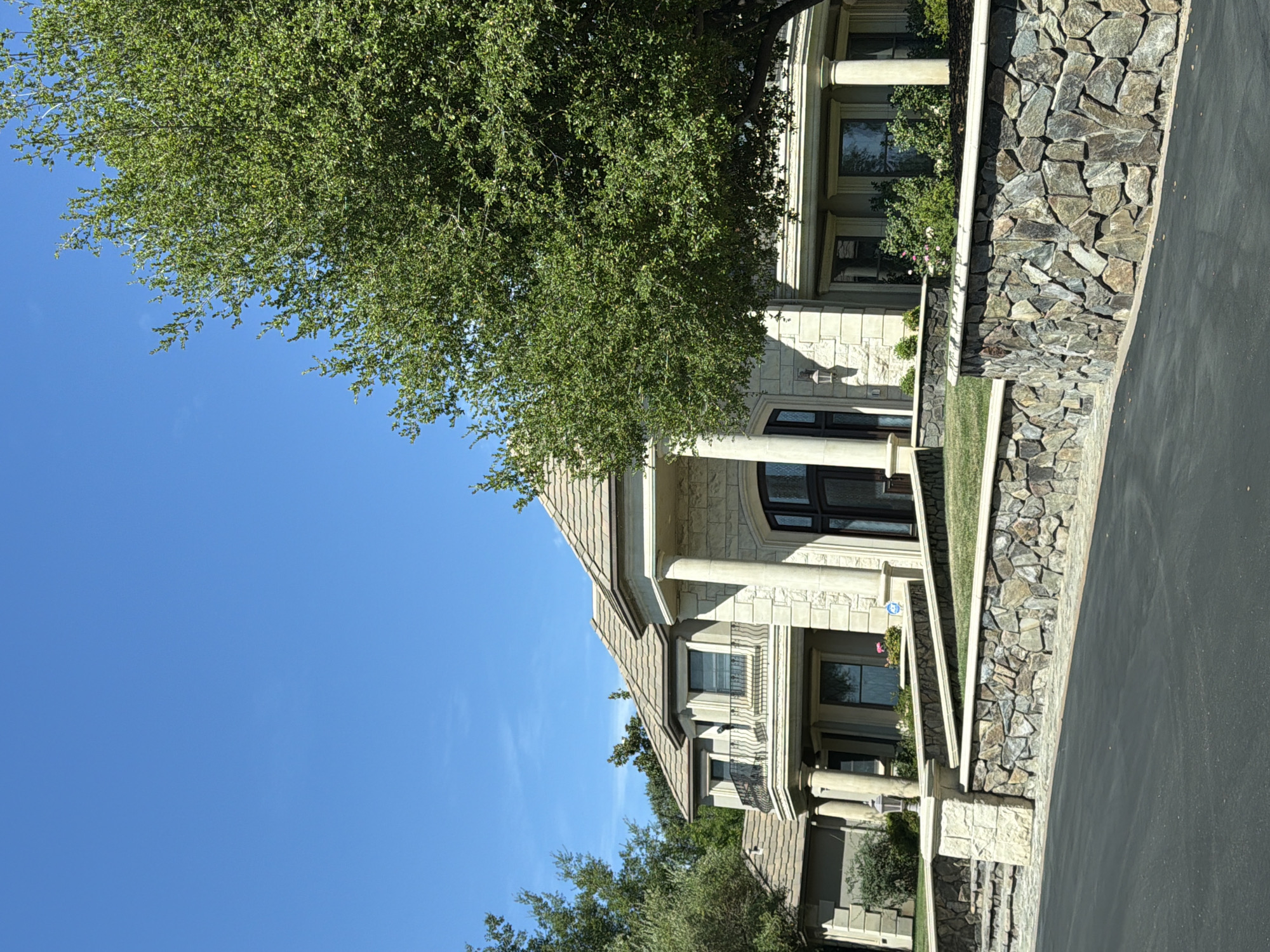 The image shows a large, upscale house with a stone facade and a tiled roof, set against a clear blue sky. A mature, leafy tree partially obscures the right side of the house. The property is fronted by a multi-tiered stone wall with landscaping and grass. A paved driveway leads up to the house. The house features large columns, arched windows, and balconies. The scene is well-lit, suggesting it was taken during daylight hours. The location is identified as Acampo, United States. There are no people or discernible actions in the image.