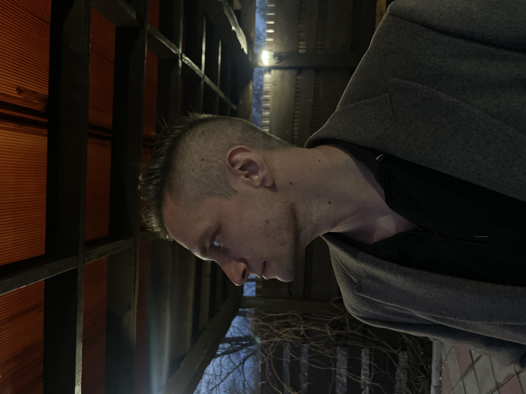 A close-up, side profile shot depicts a man, likely in his 20s or 30s, looking downwards with a neutral or contemplative expression. He has short, dark hair, possibly buzzed on the sides, and light stubble on his chin and jawline. He is wearing a dark grey, heavy-textured coat over a black zippered garment.
The background features a wooden framework of beams and slats, suggesting a sheltered outdoor area, such as a gazebo or pergola, or an indoor structure with exposed timber. Parts of this wooden structure are bathed in an orange-red glow from an unseen light source on the left, creating strong contrast with the darker, shadowed sections. In the lower right, tangled, bare branches of a tree or vine are visible against a dark, indistinct background, indicating an outdoor setting during late autumn or winter.
The scene is dimly lit, with prominent artificial light sources creating bright highlights on the man's face and reflecting off the wooden elements. The overall atmosphere is quiet, possibly at night or in the late evening, in Chaikovsky, Russia. No visible text is present in the image.
