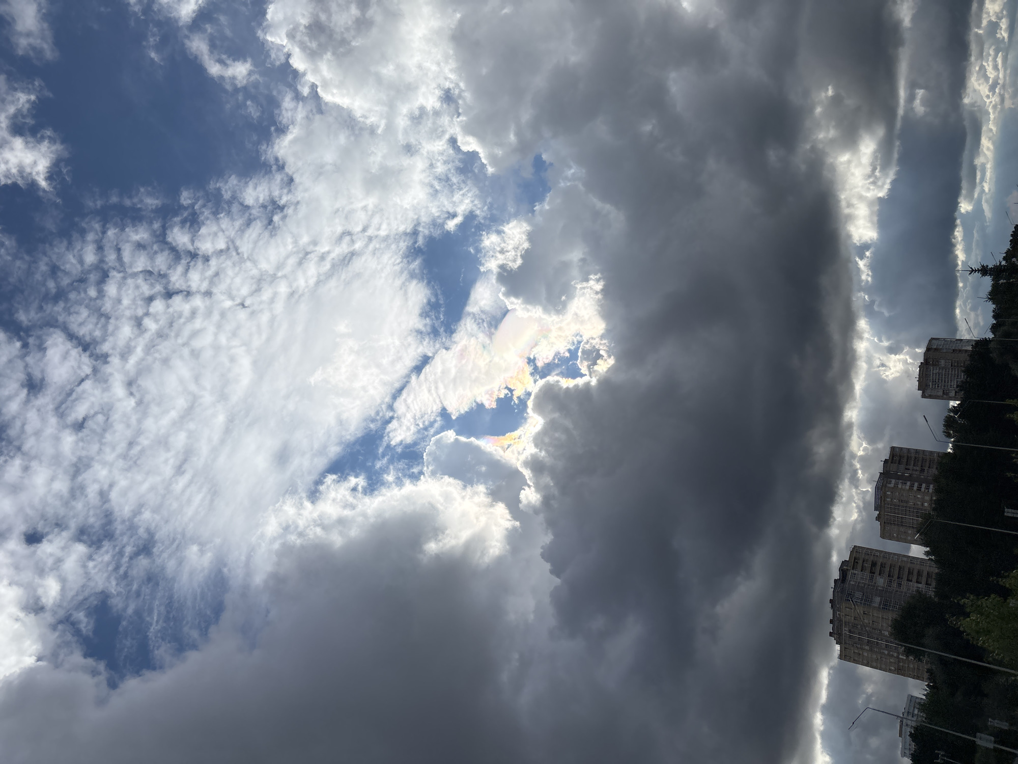 The image shows a cloudy sky over a city. In the foreground, there are dark storm clouds that partially obscure the sky. Above these, lighter clouds are visible, with patches of blue sky. A rainbow-like optical phenomenon, possibly a cloud iridescence or sun dog, is visible where sunlight refracts through ice crystals in the clouds. Below the sky, silhouetted trees and several multi-story apartment buildings are visible, suggesting an urban environment. The architecture of the buildings is consistent with residential blocks found in Russian cities. Streetlights are also visible, along with some foliage at the bottom of the frame. The overall scene suggests an overcast day with a break in the clouds allowing for the colorful atmospheric effect. Based on the architectural style of the buildings and common landscape features, this scene is likely located in Naberezhnye Chelny, Russia.