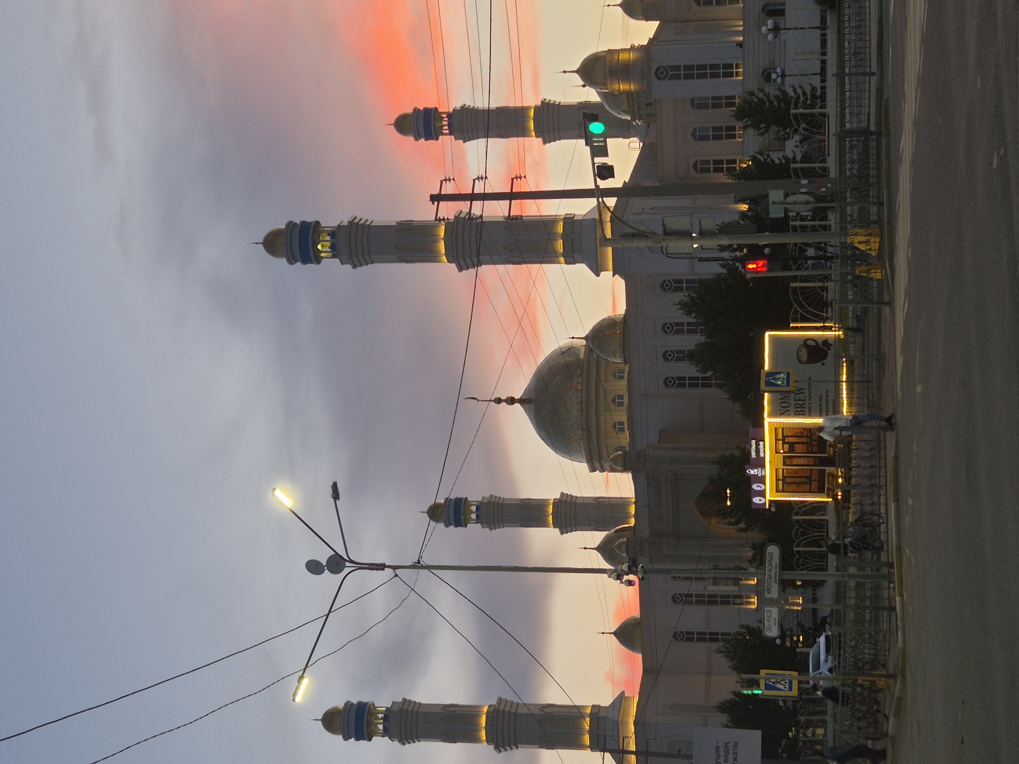 The image shows a mosque with multiple minarets and domes at sunset in Kokshetau, Kazakhstan. The sky is painted with vibrant hues of pink and orange, creating a dramatic backdrop. Power lines and streetlights are visible in the foreground, with a traffic light showing a green signal. A coffee shop with illuminated signage is located in front of the mosque. Pedestrians and a car are also present in the scene, along with bicycles parked behind a metal fence. The mosque appears to be a significant architectural landmark in the city.