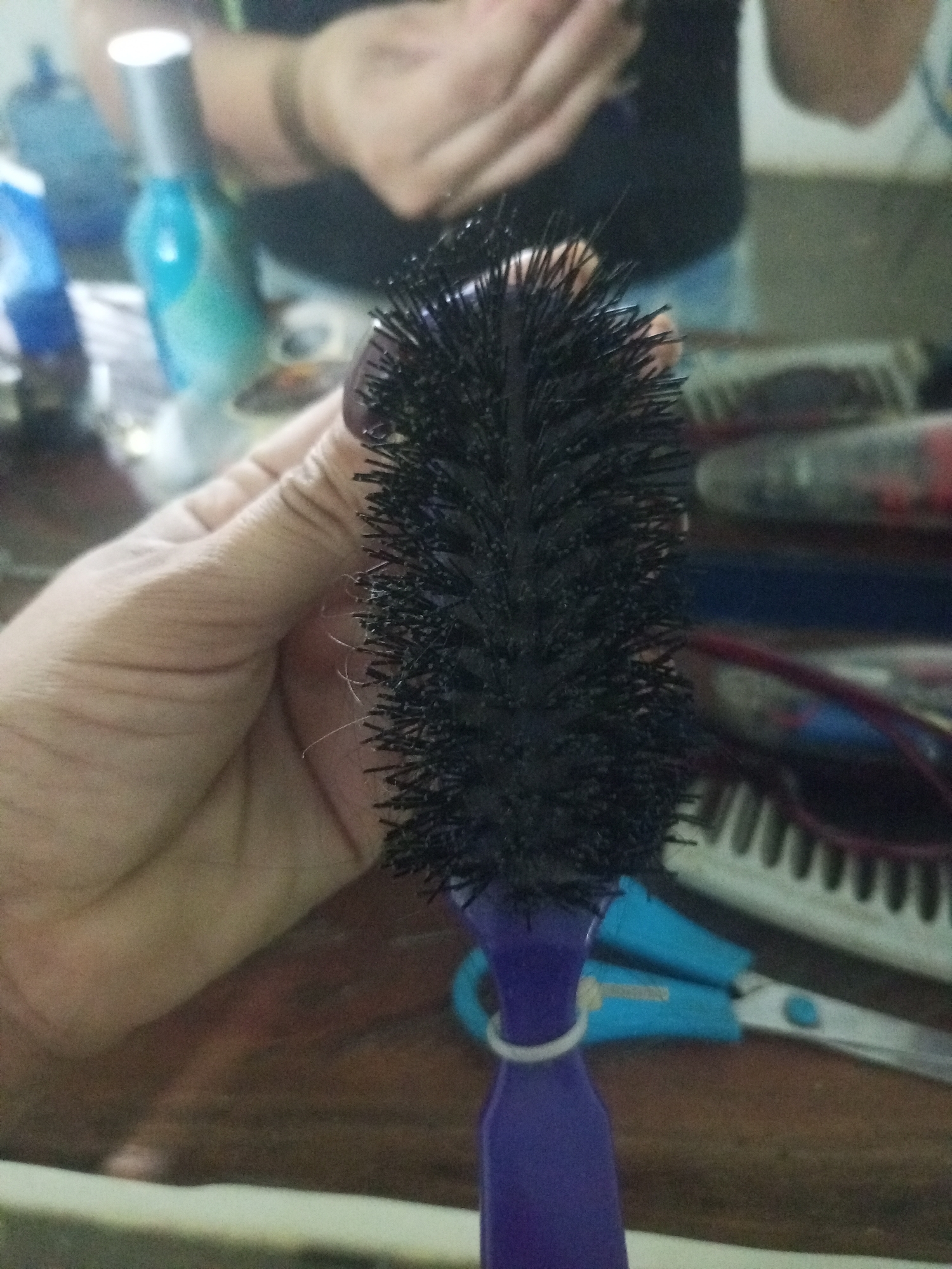 A hand is holding a black hairbrush with a purple handle. The bristles of the hairbrush are densely packed and appear slightly wet. In the background, out of focus, are various objects on a wooden surface. These include what appear to be bottles of liquid, possibly hair products, and a pair of blue scissors. The overall lighting is dim, suggesting an indoor setting. The image provides no information about the specific location within La Tagua, Venezuela.