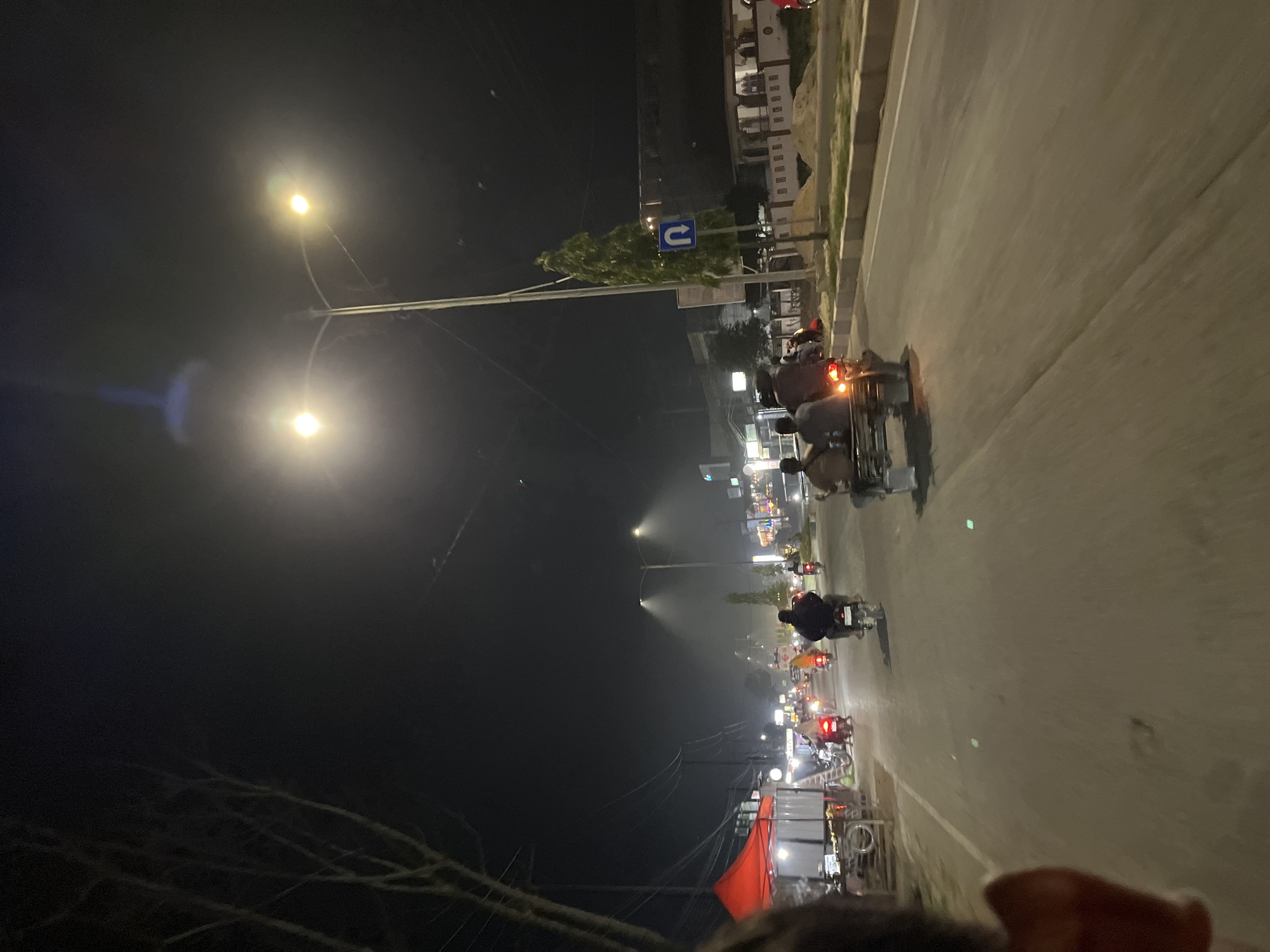 This image depicts a nighttime scene on a road in Bundar, Indonesia. The road is illuminated by streetlights, casting a warm glow on the pavement. Several motorcycles are visible, with riders and passengers heading in both directions. On the left side of the road, there's a red tent covering a stall, suggesting a market or roadside vendor. Further down the road, more stalls and buildings are illuminated, indicating an urban or semi-urban area. A tall, slender tree stands on the right side of the road, next to a U-turn sign. The sky is dark, and the overall atmosphere is one of a bustling nighttime street.