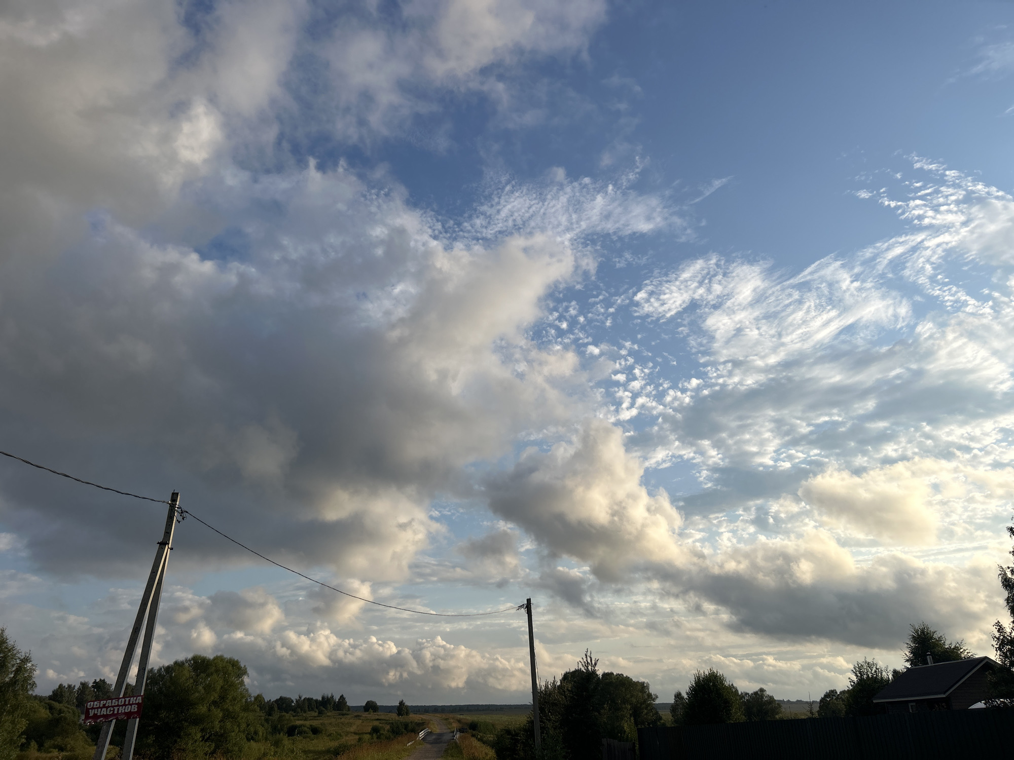 The image depicts a wide shot of a rural landscape under a dramatic sky, likely in or near Kalitsino, Russia. The foreground is dominated by a dirt road leading into the distance, flanked by grassy fields and trees. A power line stretches across the frame, supported by a concrete utility pole on the left. A red and white sign is attached to the base of the pole. In the middle ground, more trees and vegetation are visible, and a dark wooden fence runs along the right side of the frame, partially obscuring a small, dark-roofed building. The sky is a significant element of the image, filled with a mixture of dark, heavy clouds and wispy, lighter clouds, suggesting changing weather conditions. The overall impression is one of a serene but perhaps slightly moody rural setting.