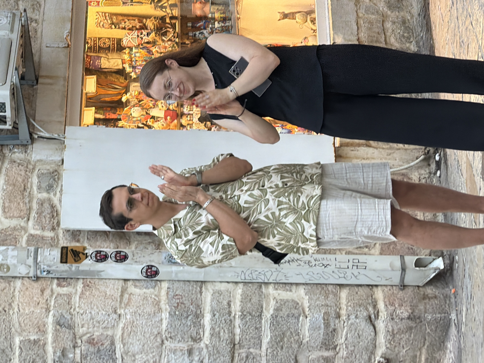 In Budva, Montenegro, on a cobblestone street, a man and a woman stand side-by-side, clapping. The man, wearing sunglasses, a short-sleeved button-up shirt with a palm leaf pattern, and light-colored shorts, has his hands clasped in front of him. A black bag hangs from his left hip. The woman, to his right, wears a black sleeveless top and wide-legged black pants. She has glasses and her hands are also clasped. She holds a smartphone in her right hand. They are positioned in front of a shop window filled with various colorful trinkets and souvenirs. A stone wall with graffiti and a downspout are visible to the left of the pair. The scene appears to be outdoors in an urban setting.