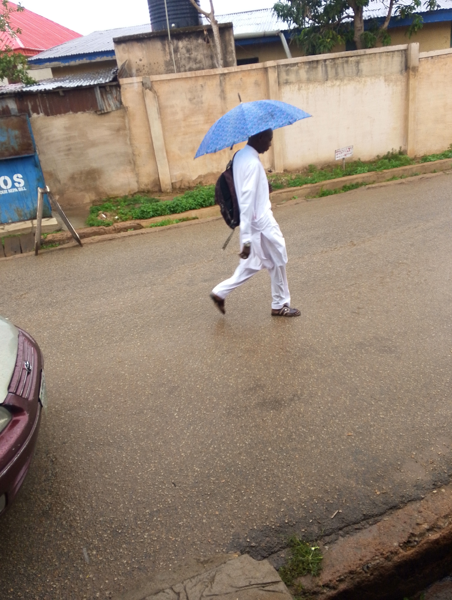 A person walks along a wet street in Jos, Nigeria. The person is wearing a white outfit, a backpack, and sandals, and is holding a blue umbrella. A car is parked on the left side of the image. Behind the person is a high wall. Some greenery is visible on both sides of the street. Buildings with corrugated iron roofs are visible in the background.