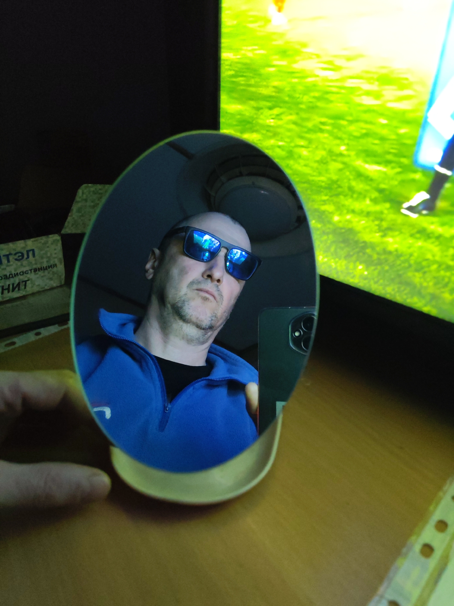 An indoor medium close-up shot captures a man taking a selfie by reflecting his smartphone in a small, oval-shaped mirror. The man, who is bald with some stubble, is wearing dark blue reflective sunglasses and a blue zip-up fleece jacket over a black t-shirt. He holds the mirror with his left hand, while his right hand holds a dark-colored smartphone, whose prominent rear camera module is visible in the mirror's reflection, pointing towards his face.
In the background, to the right of the mirror, a large screen or television displays a vibrant green expanse, reminiscent of a sports field or video game environment, with a partial figure in blue and white visible on the right edge. To the left of the mirror, also in reflection, there's a dark background with some lighter objects, possibly boxes or documents. Visible text on one of these reflected objects appears in Russian: "ИТЭЛ", "радиостанции", and "НИТ".
The mirror, resting on a light-colored base, is placed on a wooden desk or table. Part of a document with punched holes is visible on the far right of the desk. The lighting in the scene appears to be artificial, casting a cool tone, particularly within the mirror's reflection. The man's expression behind his sunglasses seems neutral or contemplative.