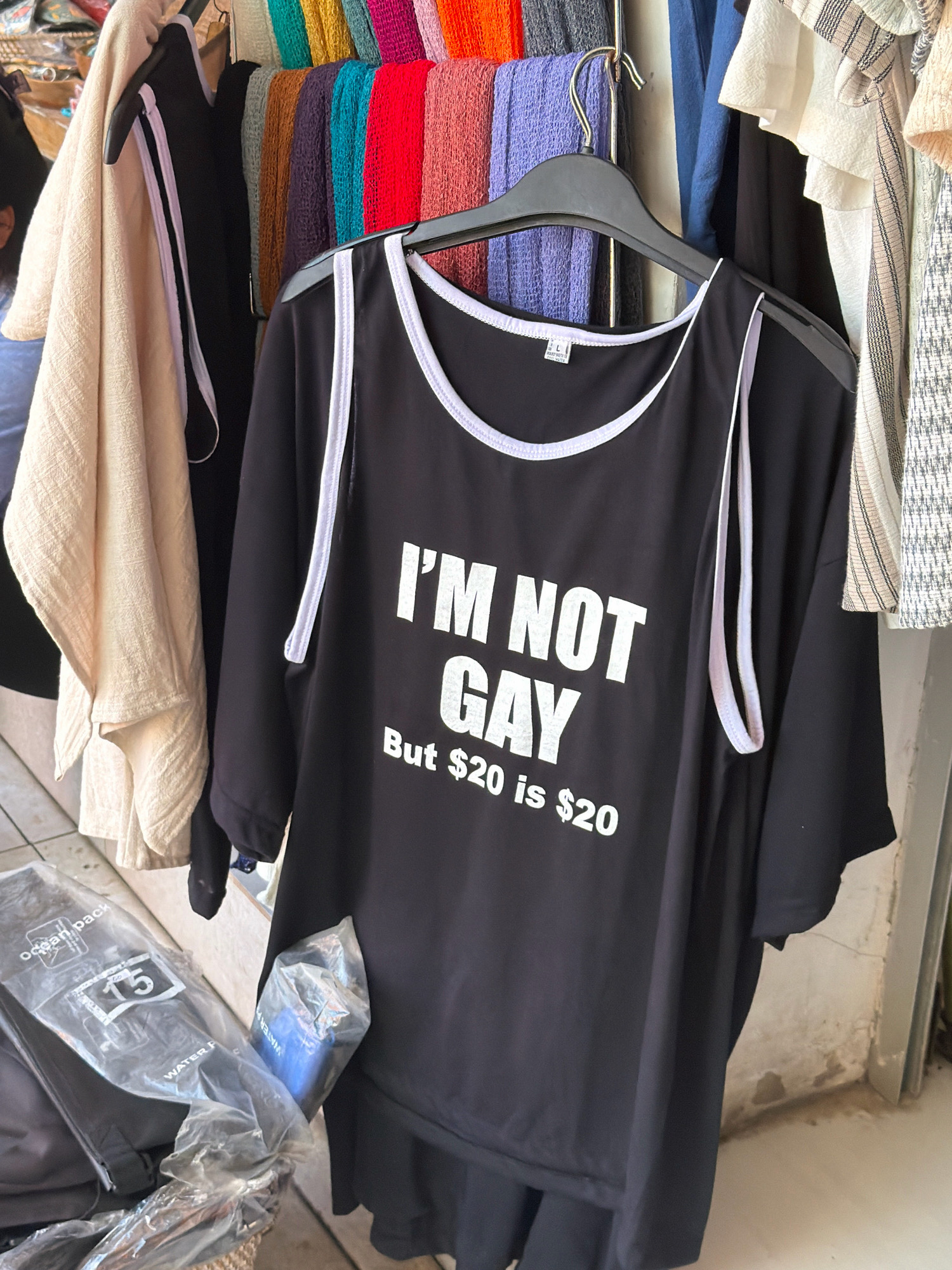 In North Kuta, Indonesia, a black t-shirt with white lettering is displayed on a hanger in a market or shop. The t-shirt reads "I'm NOT GAY But $20 is $20". Behind the t-shirt, various colored textiles and other clothing items are visible, hanging on racks. To the left of the t-shirt, a beige garment is draped. In the foreground, clear plastic packaging with some text and a white number "15" is visible. The background suggests an indoor or covered outdoor market setting with a concrete floor and a wall.