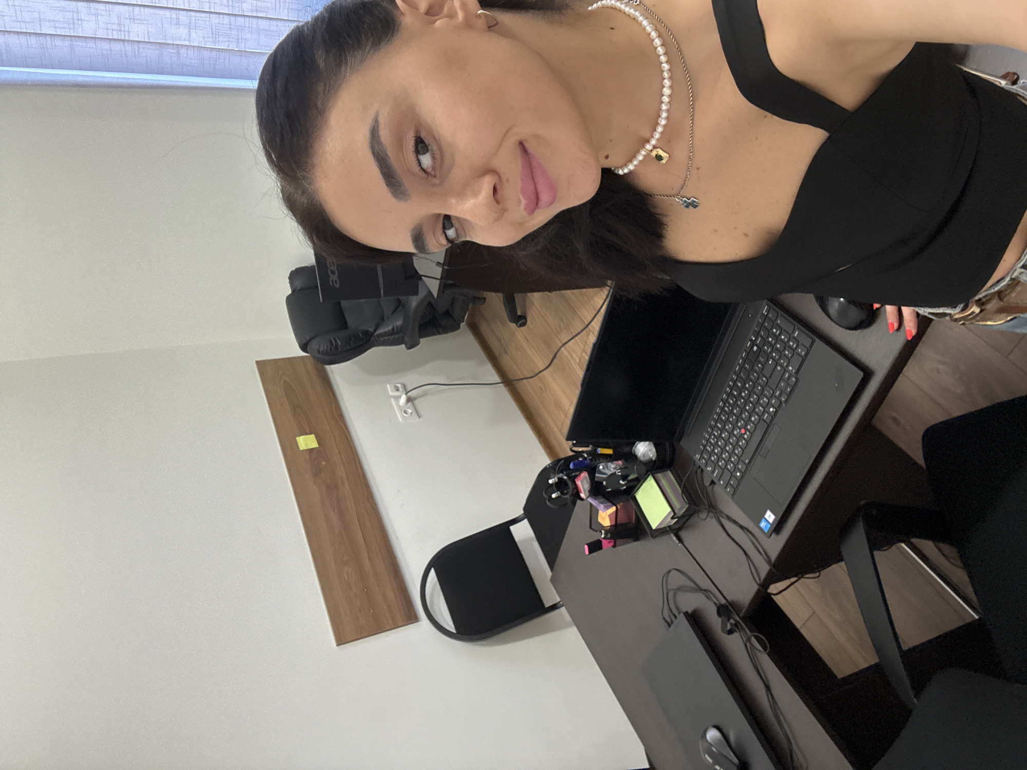 The image shows a woman in a black top and jeans, wearing a pearl necklace, in what appears to be an office setting. She is looking directly at the camera with a playful expression, her tongue sticking out slightly. A laptop, various office supplies, and a wooden plank are visible on a desk. A black office chair is also in the frame, along with a white wall and a window with blinds. The overall impression is of a casual, perhaps slightly messy, workspace. The lighting suggests it might be daytime, and there are no obvious weather cues. There is text visible on the laptop, but it is not clearly legible. The scene appears to be indoors, possibly in an office or home office environment.