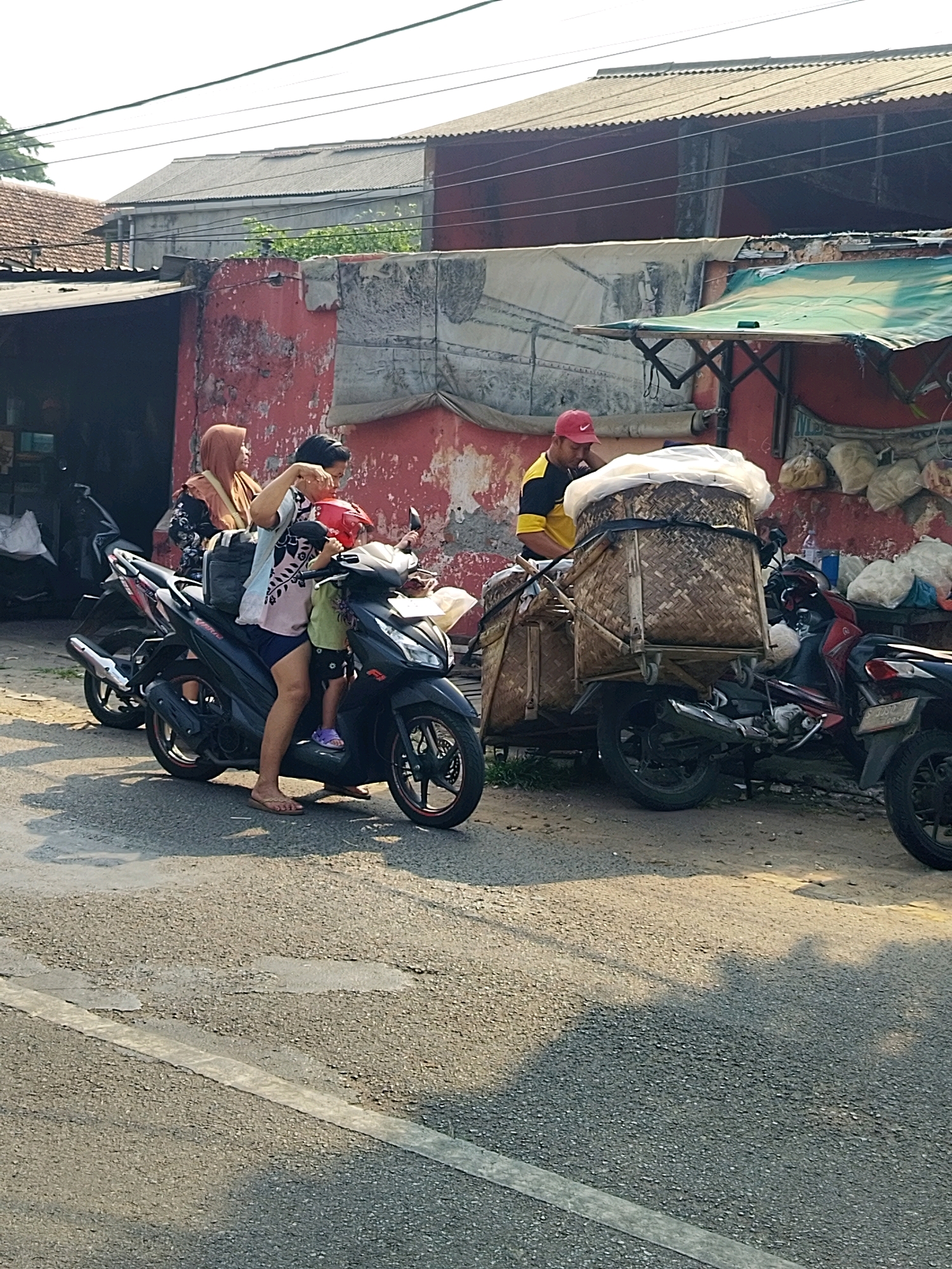 In Mojokerto, Indonesia, a family is on a motorcycle. The mother, wearing a headscarf, is sitting behind the driver, who is holding a child. The child is wearing a red helmet. The motorcycle is black and appears to be a scooter. In the background, there is a building with a red wall and a corrugated iron roof. There is also a market stall with a green awning. The scene appears to be on a street or road.