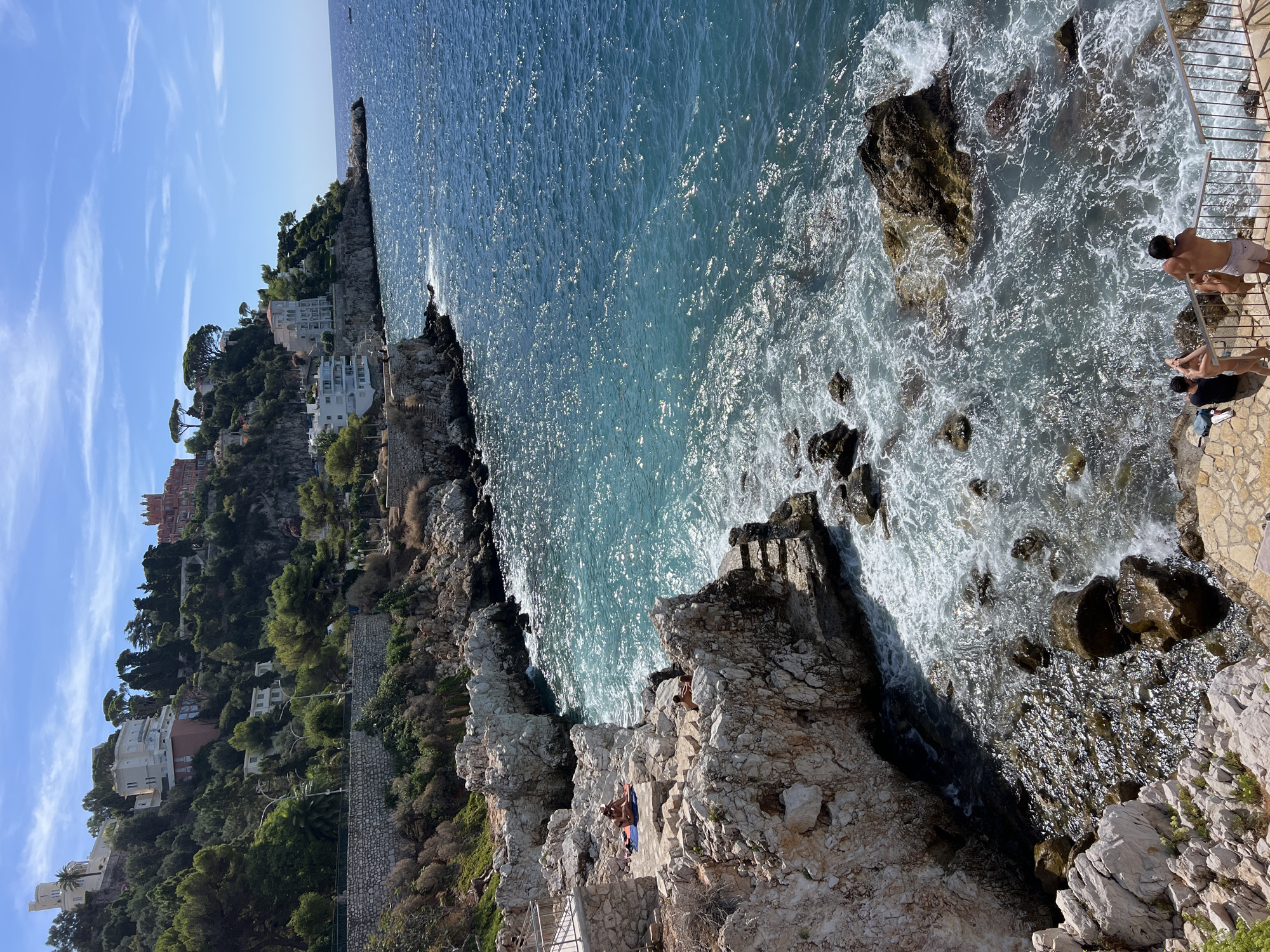 This image depicts a rocky coastline in Nice, France, with the Mediterranean Sea in the foreground and affluent buildings dotting the hillside in the background. The sea is a vibrant turquoise, with waves gently crashing against the rocks. Several people are visible on the rocks, some sunbathing and others wading into the water. A stone staircase leads down to the water's edge. The hillside is covered in lush green vegetation and dotted with elegant villas and apartment buildings, hinting at a luxurious coastal setting. The sky is a clear blue with a few wispy clouds.