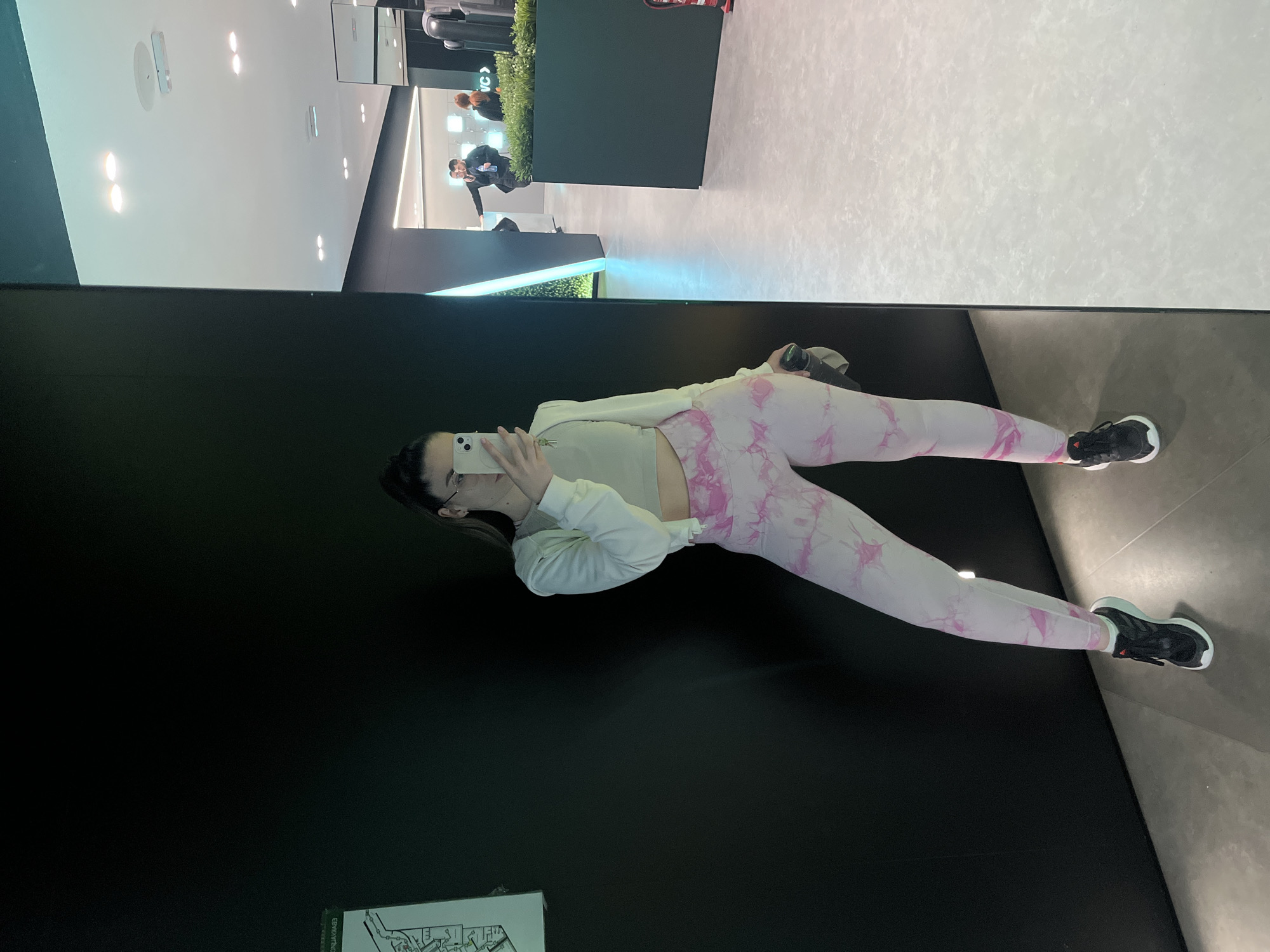A full shot captures a woman reflected in a mirror. She is dressed in athletic wear: a white jacket, a cropped top, and pink patterned leggings, with black sneakers. She is holding a smartphone, ostensibly taking a selfie. Behind her, a black wall dominates. The mirror reflects a brightly lit modern interior with a white ceiling, recessed lighting, and hints of other individuals in the background. It is not possible to tell the specific city of Unknown.