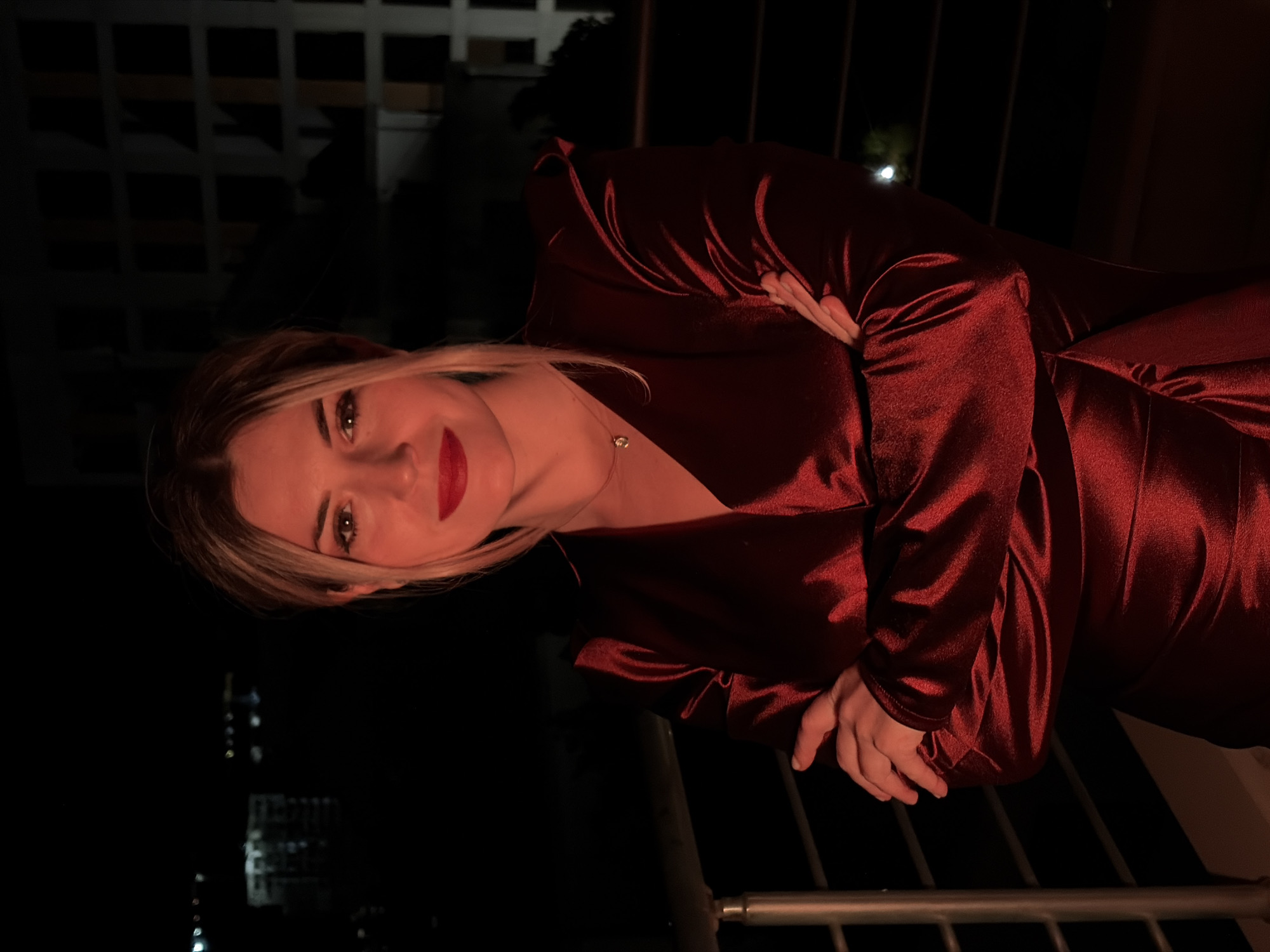 A blonde woman with red lipstick and a dark red satin dress stands with her arms crossed on a balcony at night. The background is dark with some faint lights from buildings and structures in the distance, suggesting an urban environment. The woman is looking directly at the camera with a neutral expression. A railing is visible in the foreground. The setting is likely a high-rise building or apartment complex in the city of Budva, Montenegro, given the visible urban lights and the presence of a balcony.