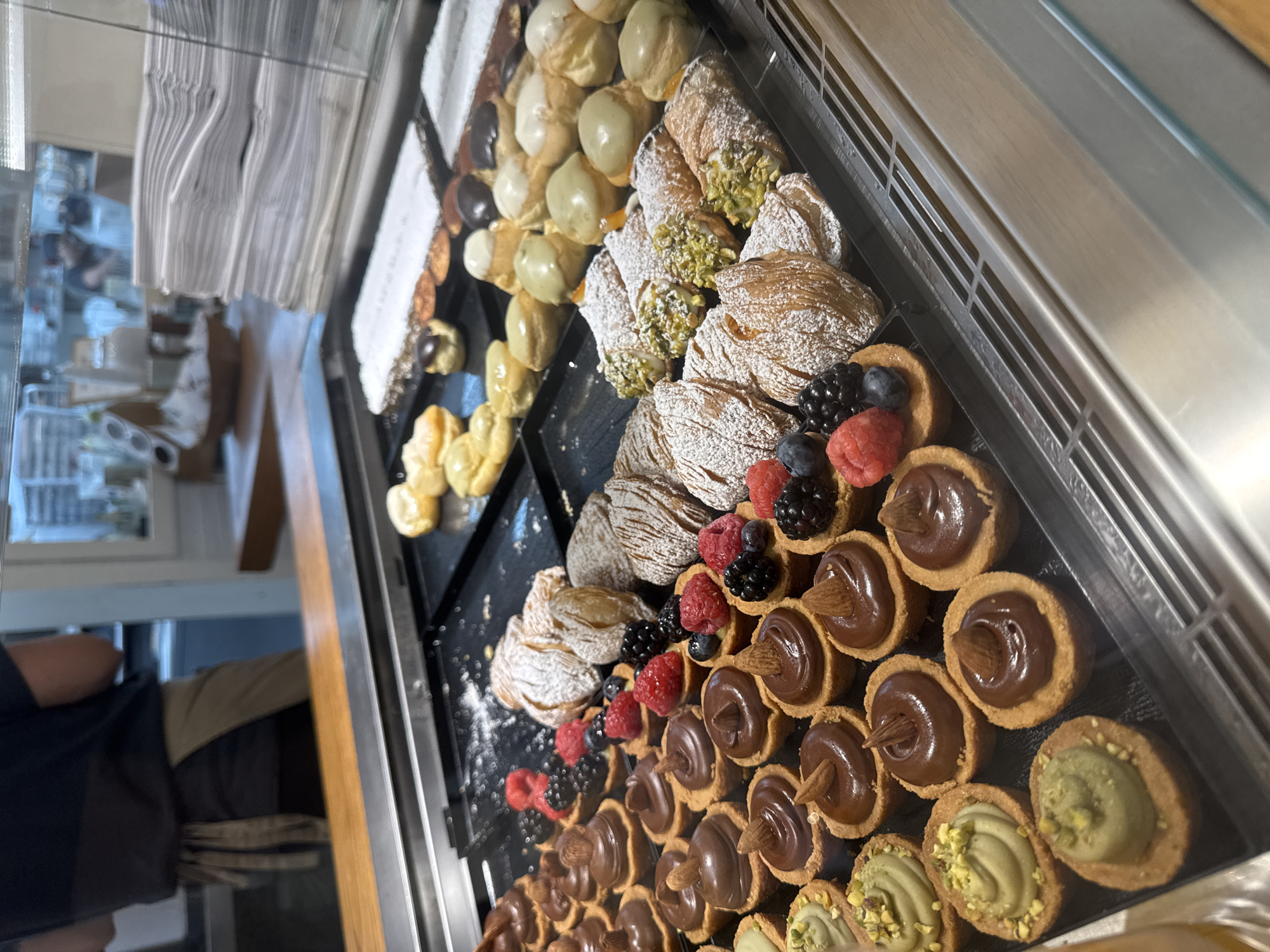 The image depicts a display case filled with various pastries and desserts, likely in a bakery or dessert shop. The pastries include cannoli filled with cream, Sfogliatella with powdered sugar, cream puffs, and small tarts filled with chocolate and topped with almonds or pistachio cream. Some of the tarts are decorated with fresh berries, including raspberries and blueberries. The pastries are arranged neatly on black trays inside the display case. The location is Aventura, United States of America. A person wearing a black shirt and tan apron can be seen partially in the reflection of the display case.