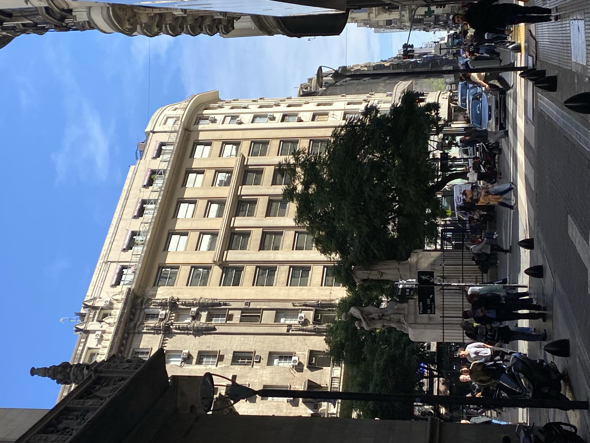 The image depicts a street scene in Buenos Aires, Argentina. A tall, light-colored building occupies the left side of the frame. The building's architecture features repetitive windows, ornate details, and multiple air conditioning units. A green tree partially obscures the building.
On the right side of the frame, a group of people stand on the sidewalk near a street with cars and a crosswalk. The people appear to be waiting to cross the street. The street and sidewalk are separated by black bollards and a fence. A sign with "Florida" and numbers "50" and "100" is visible. Pedestrians and some motorcycles are also seen.
The sky is blue with some clouds. The image is taken from a low angle, looking up at the building. Elements of another building partially block the bottom left corner of the frame.