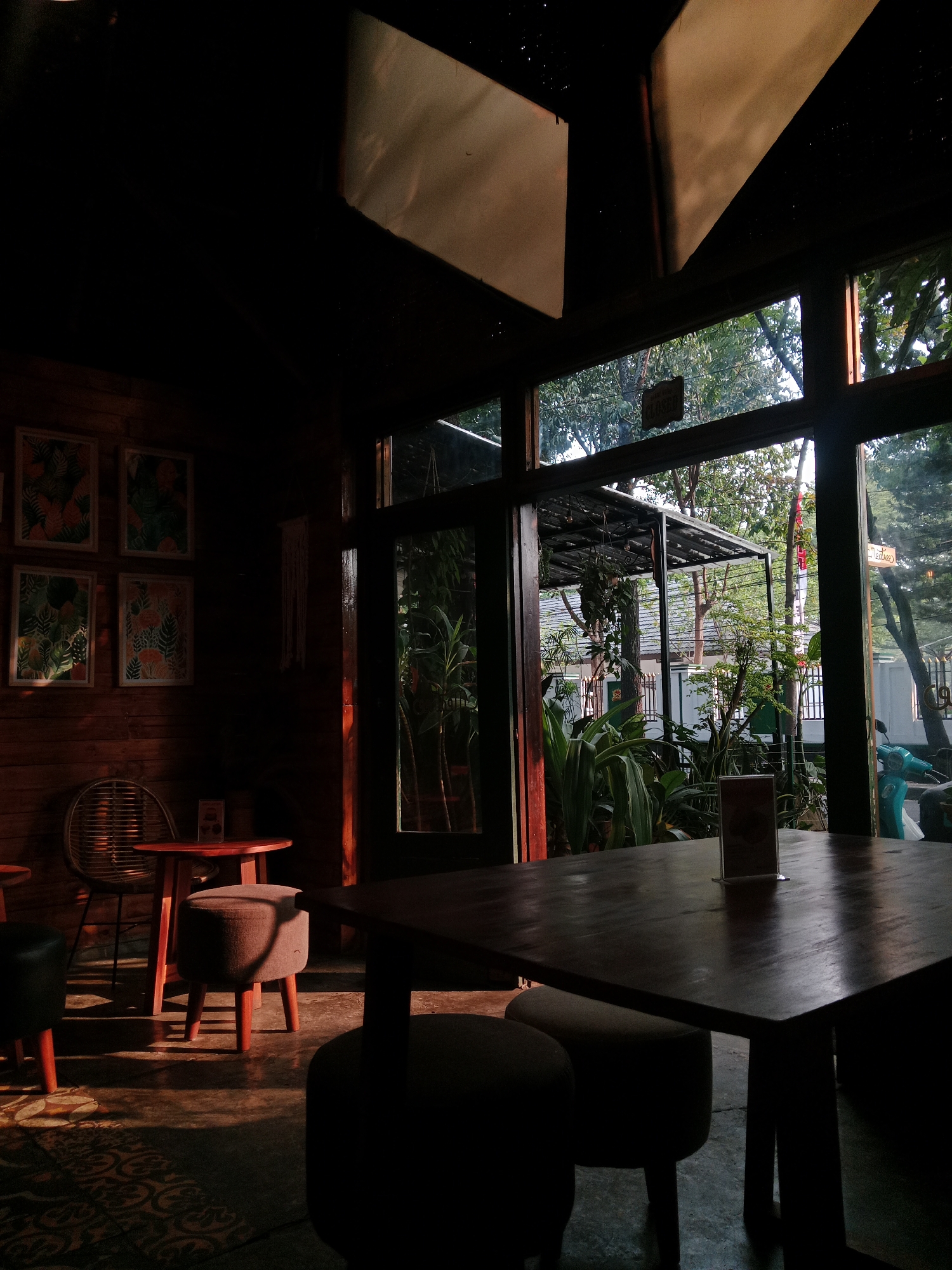 This is an interior shot of a cafe with large glass windows looking out onto a garden area. The cafe has wooden walls and a dark ceiling with some lighting fixtures. There are several tables and chairs visible, including a large wooden table in the foreground with two stools. A smaller table with a chair and a fan is positioned against the left wall. Four framed pictures of plants hang on the wooden wall. The sunlight streaming through the windows creates strong shadows across the floor and furniture. The outdoor area appears lush with plants and trees, and some structures are visible, including a covered patio area. A turquoise motorcycle is parked outside to the right. The image has a warm, golden hour lighting aesthetic, typical of cafes in Bandung City, Indonesia, which often feature outdoor or garden seating to enjoy the tropical climate. The overall ambiance suggests a relaxed and dimly lit environment.