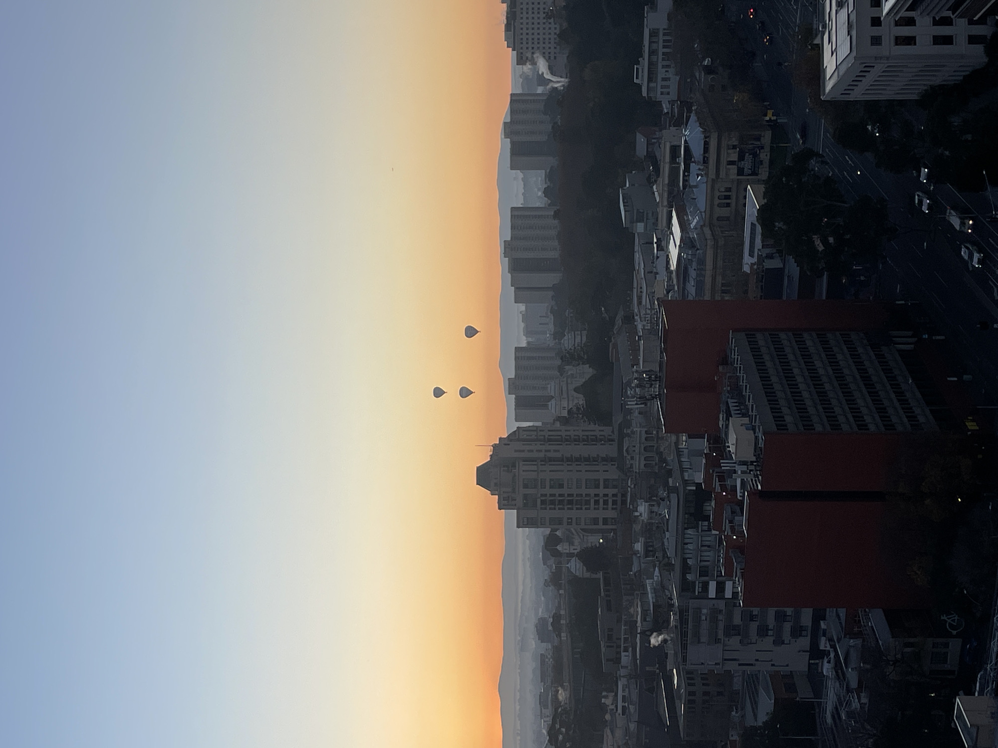 An aerial view captured at dawn showcases the city of Melbourne, Australia, with a blend of modern and older architecture. Four hot air balloons are visible in the sky, three of which are clustered together, while the fourth is further to the left. The sky displays a gradient of colors, transitioning from a soft blue at the top to a warm orange at the horizon, indicating sunrise. In the distance, misty hills are faintly visible. The urban landscape is characterized by a mix of multi-story buildings, some appearing residential and others commercial, interspersed with trees. A prominent, tall building with a textured facade stands out in the mid-ground. Smoke or steam is rising from a few buildings, suggesting early morning activity. Streets with vehicles are visible in the lower portion of the image, and the lighting suggests it is either early morning or late evening.