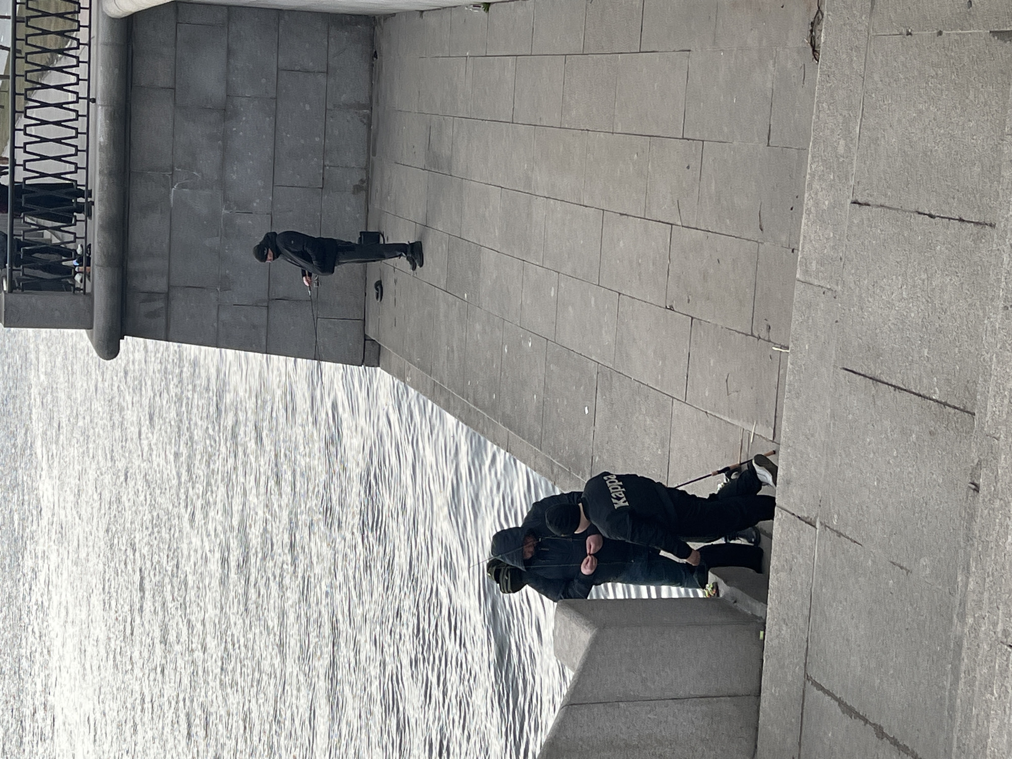 The image captures a scene on a gray stone embankment in Saint Petersburg, Russia, adjacent to the Neva River. Two individuals are seen fishing over the edge of the embankment, both dressed in dark clothing and hats, with one wearing Kappa-branded attire. A third person stands further along the embankment, also dressed in black, holding a fishing rod. A section of decorative black iron railing is visible on the left side of the image. The Neva River is in the background, reflecting sunlight.