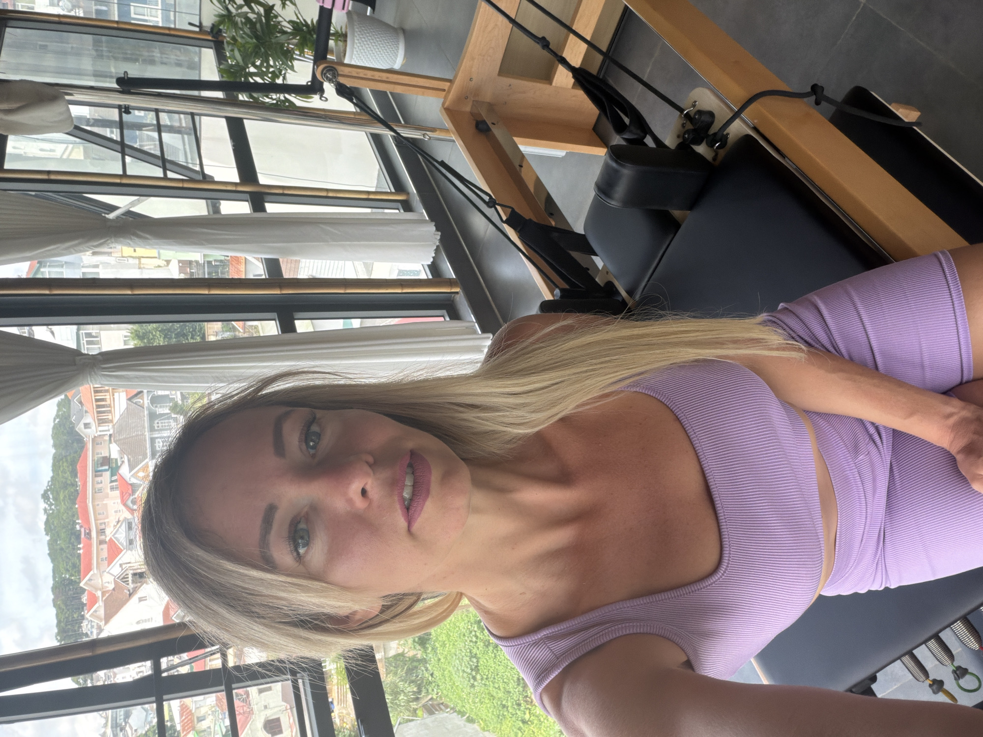 A woman with blonde hair is taking a selfie in a room with a large window overlooking a city with red-tiled roofs. She is wearing a purple athletic set. In the foreground, there is a piece of exercise equipment, possibly a Pilates reformer. The room appears to be a fitness studio, with bamboo accents on the window frames and a potted plant in the background. The city visible outside the window is characteristic of Da Lat, Vietnam, known for its French colonial architecture and hilly terrain.