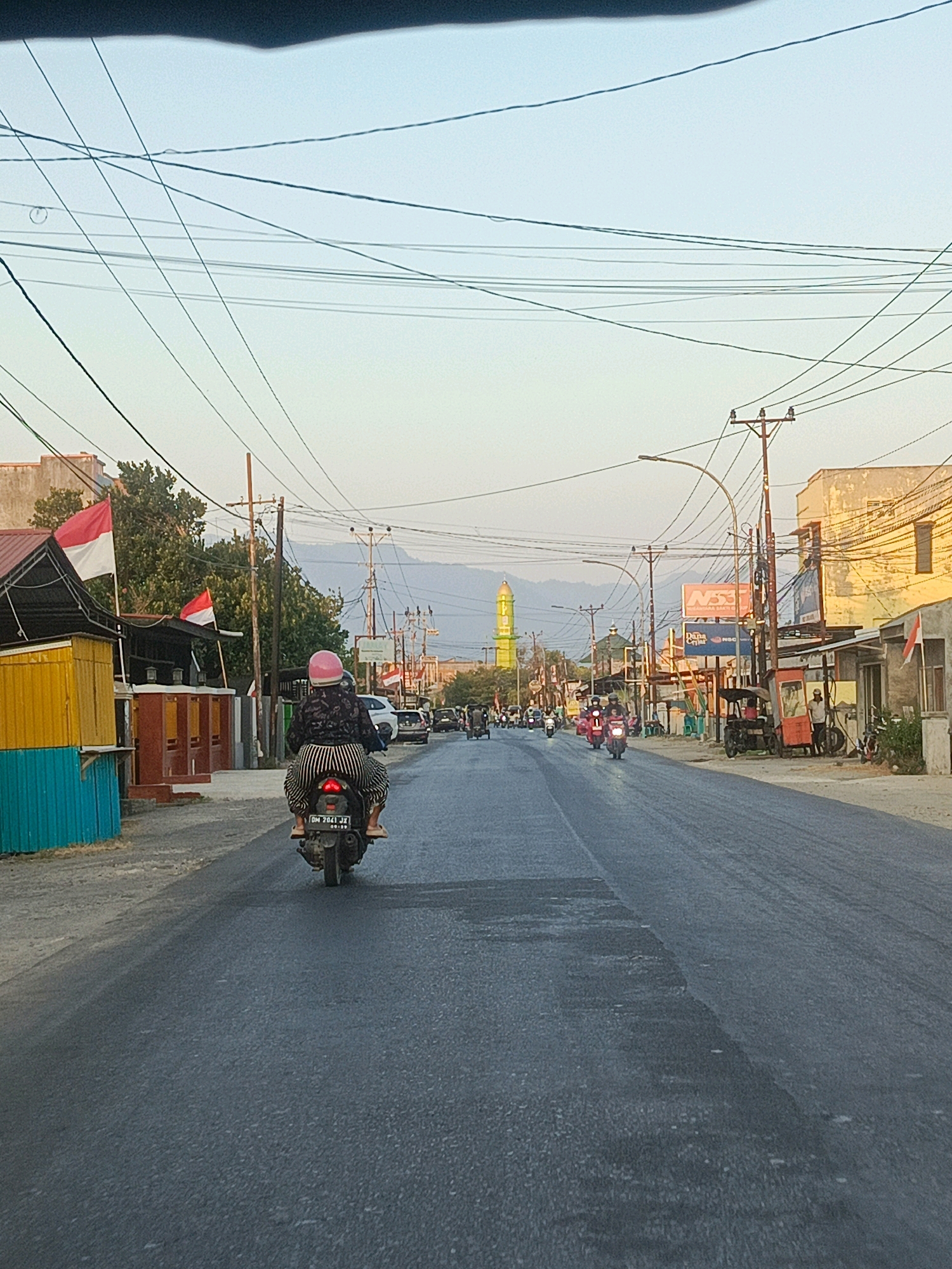 A side-view, rear-facing, medium-shot captures a person riding a black scooter with a black and white patterned shirt and striped pants, wearing a pink helmet, down a paved road in Gorontalo, Indonesia. The rider's scooter has a license plate reading "DM 2041 JX". The road is lined with buildings on both sides. On the left, a yellow and turquoise structure with red accents flies two Indonesian flags. Across the street, various businesses, including one with a red and white sign reading "NSS", are visible, along with more Indonesian flags. Several other scooters and vehicles are present on the road, some moving and some parked. In the distance, a green mosque with a gold dome stands out against a hazy sky and mountains. Overhead, a complex network of power lines crisscrosses the frame. The lighting suggests it is late afternoon or early morning, with soft, diffused light casting long shadows.