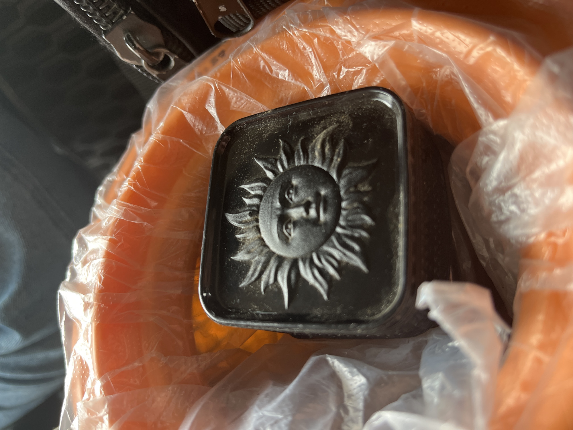 The image presents a close-up view of a dark, square-shaped object featuring a prominent, raised relief of a sun. The sun design includes a human-like face at its center, surrounded by numerous radiating rays. Fine particles, possibly dust or debris, are visible on the object's surface, particularly around the intricate sun design. The object appears to be made of a solid, heavy material, possibly metal or resin.
This sun-emblazoned item is partially resting on or wrapped within a transparent, crinkled plastic bag. Beneath the plastic bag, a vibrant orange object provides a striking color contrast against the dark sun piece and the translucent plastic. In the upper left corner, a portion of a dark zipper with a metallic pull tab is visible, suggesting the presence of a bag or another item with a fastening mechanism.
The lighting is bright, creating distinct highlights on the plastic bag and the raised contours of the sun. The scene depicts a static arrangement of these objects, without any discernible activity or event in progress. No text is visible on any of the items. The time of day, weather conditions, or specific location cannot be determined from the image.