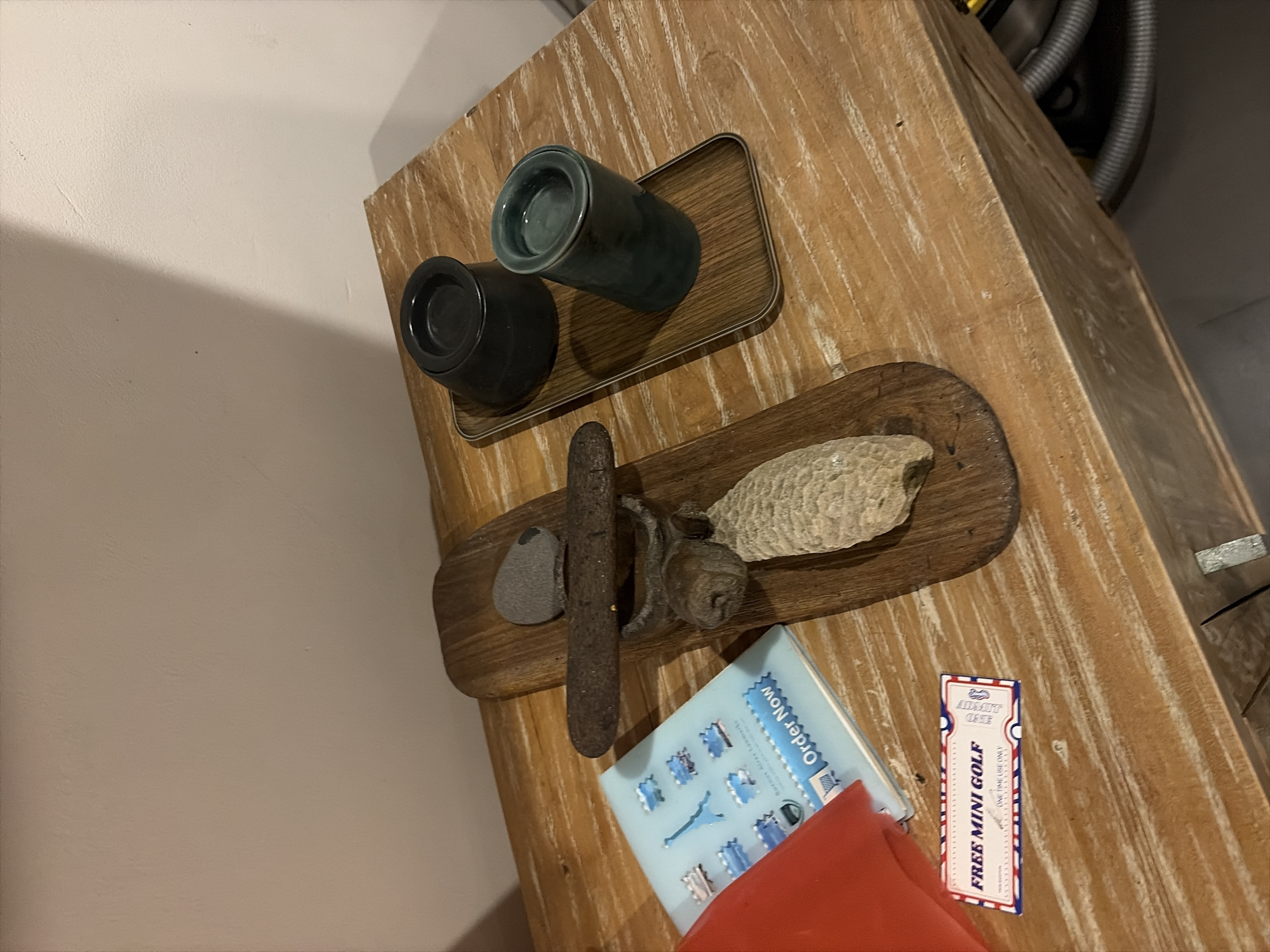 A close-up, high-angle shot shows a rustic wooden table with various objects. On the left, a light blue booklet with images of Paris landmarks, including the Eiffel Tower, is visible, along with a folded red object. Below the booklet is a small white and red ticket that reads "FREE MINI GOLF." To the right of these items is a wooden sculpture made of a long piece of wood, a grey stone, and a corn cob. The sculpture is mounted on a larger piece of wood. Further to the right, a rectangular tray holds two dark ceramic containers, one black and one teal. The background consists of a light-colored wall with a shadow cast from an unseen light source. The table's surface has prominent wood grain and appears to be part of a dresser or cabinet, with a drawer pull visible at the bottom. The scene is indoors, and no specific location in Mengwi, Indonesia, is identifiable from the visual cues.