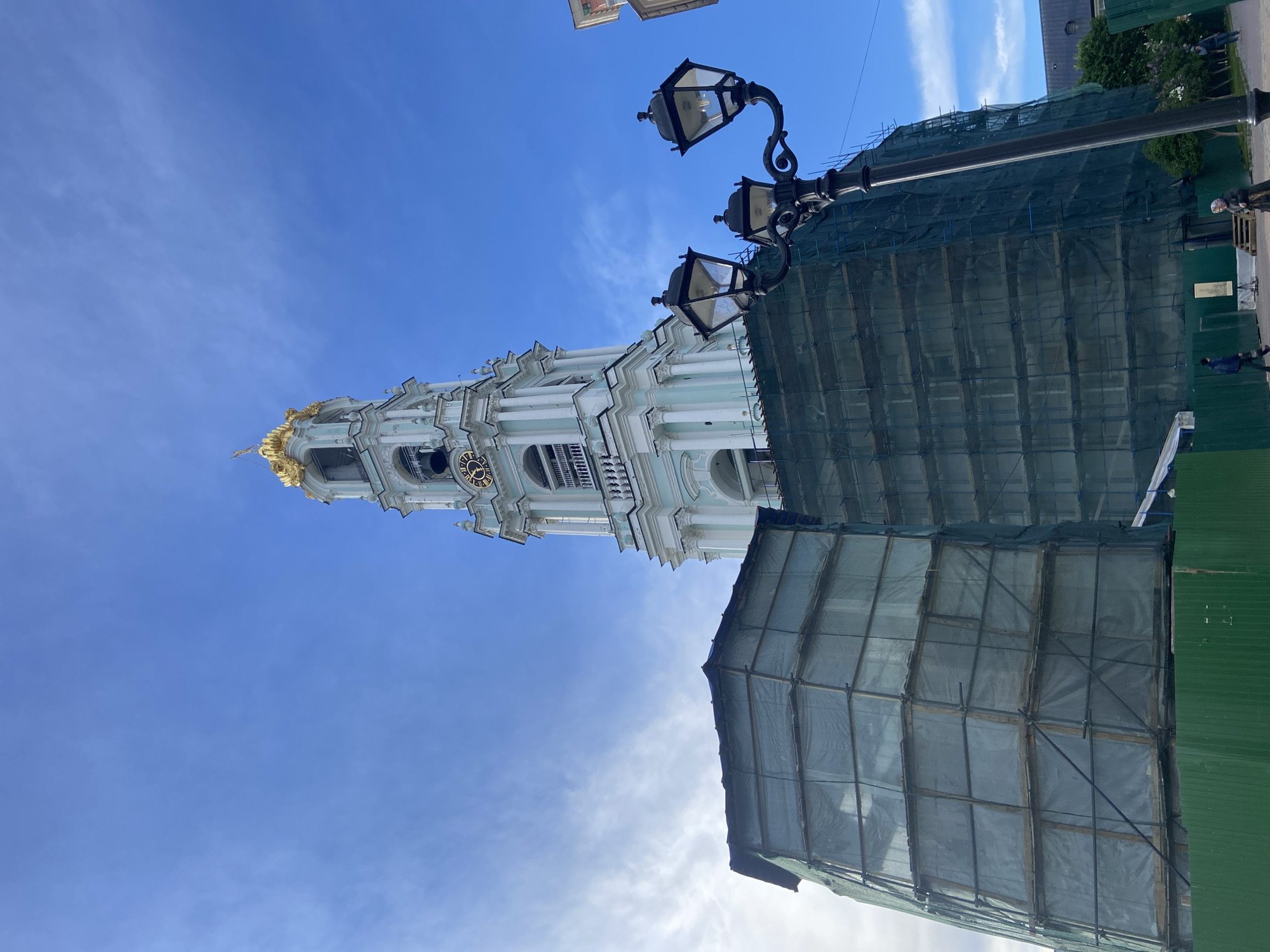 The image depicts a view of the Sergiev Posad Lavra monastery complex in Sergiyev Posad, Russia. A tall, light-colored bell tower with a gold dome is visible against a clear blue sky with some wispy clouds. Part of the monastery is covered in scaffolding with green netting. To the right of the tower, there is a lamppost with multiple lanterns. Some people can be seen in the background on the right side of the image, near a green fence.