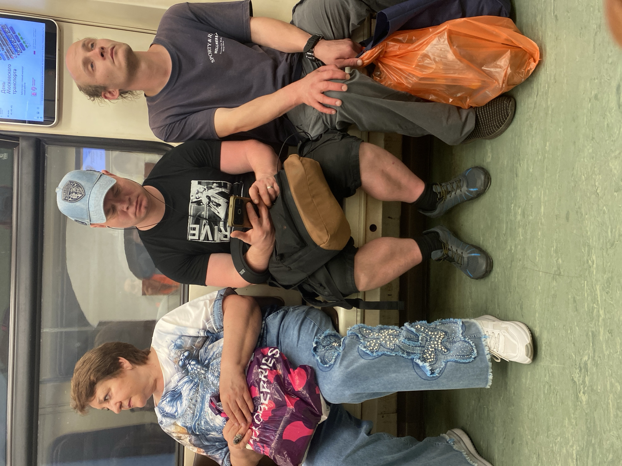 Three people are seated on a subway car in Moscow, Russia. On the left is a woman with short brown hair, wearing a white and blue patterned shirt and blue jeans adorned with a decorative applique on the right leg. She holds a purple shopping bag with pink circles in her lap. In the center, a man wearing a blue baseball cap, black t-shirt, and black shorts is looking down at his phone, which he holds in his right hand. Beside him, on the right, a man with short, thinning hair is wearing a grey t-shirt and grey pants. He has a black watch on his left wrist and is holding an orange plastic bag. All three are seated next to each other on a bench. The floor of the subway car is a light green color.