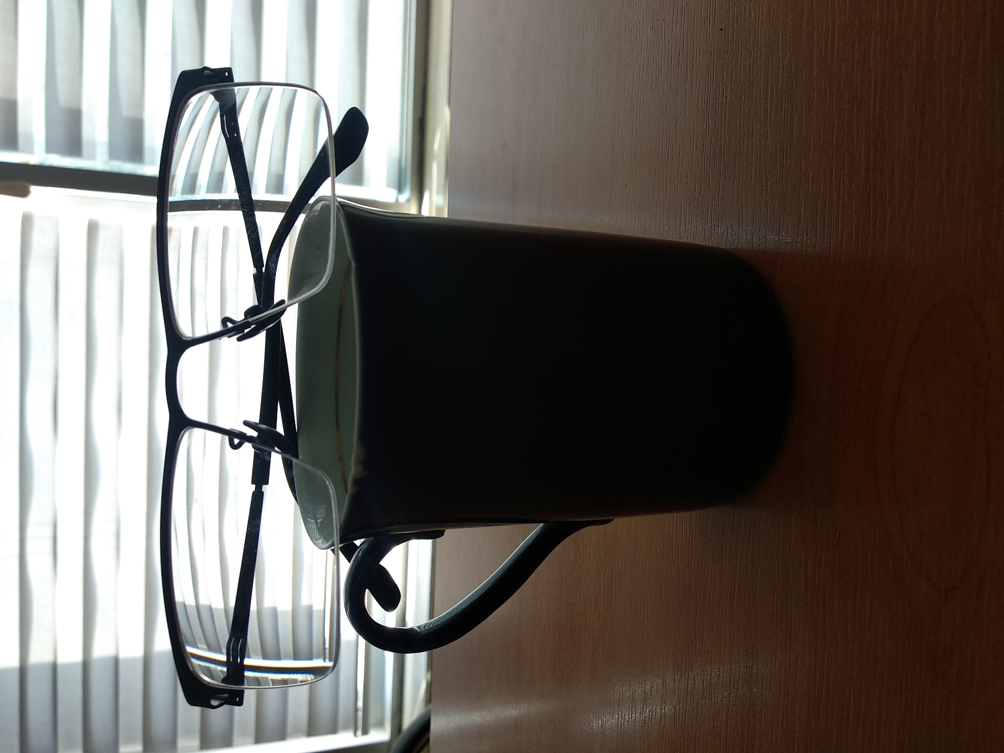 A pair of black-rimmed eyeglasses rests on top of a dark-colored mug, which is positioned on a wooden surface. The mug has a curved handle visible on the left side. In the background, a window with horizontal blinds is illuminated by light. The scene appears to be indoors, likely on a desk or table.