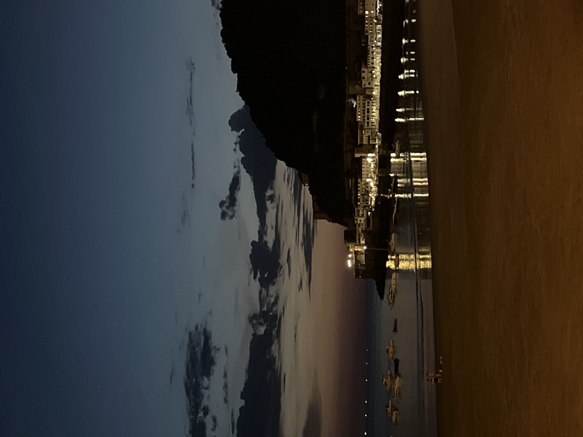The image depicts a twilight scene at La Concha beach in San Sebastián, Spain. The sky is a deep blue, fading to a pale orange near the horizon, with scattered dark clouds. Two distant, bright stars are visible in the upper sky. Below the horizon, the lights of a coastal town twinkle, illuminating a rocky shoreline and buildings that line the bay.
The water is calm, reflecting the town's lights. Several small boats are anchored in the bay. In the foreground, a sandy beach stretches across the bottom of the frame. Two individuals, a man and a woman, are standing near the water's edge, facing away from the camera. They appear to be engaged in a conversation or observing the scene. The overall atmosphere is peaceful and serene, capturing the transition from day to night in a picturesque coastal setting.