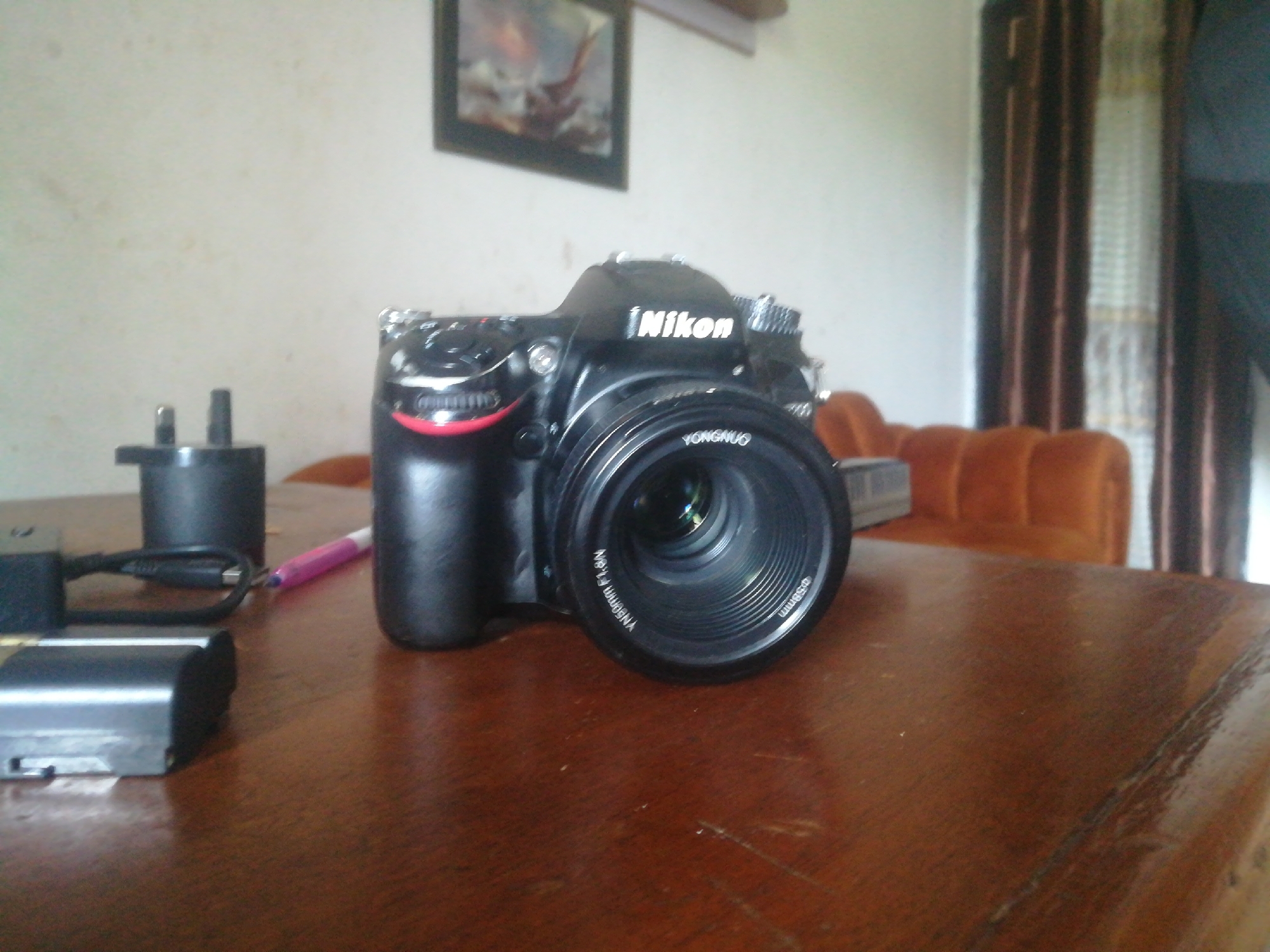 A black Nikon DSLR camera with a lens is centered on a polished wooden table. To the left of the camera are a black power adapter and a silver-colored battery. A pink pen lies on the table near the power adapter. In the background, a framed picture hangs on a light-colored wall. A glimpse of an orange upholstered chair and vertical blinds are visible through the doorway on the right. The overall scene suggests an indoor setting, likely a room in a home or office.
