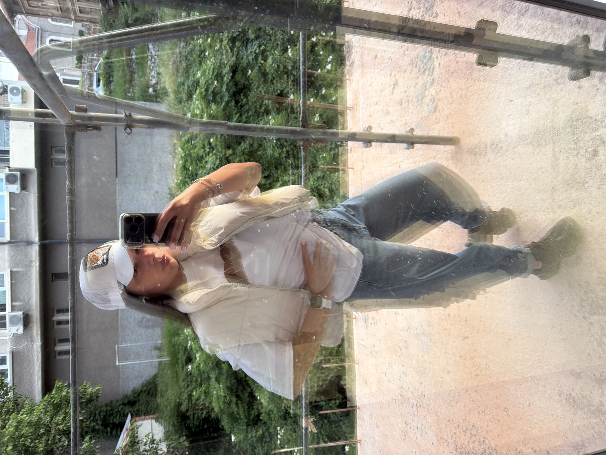 The image shows a young woman standing on a balcony, taking a selfie in a mirror. She is wearing a white baseball cap with an embroidered patch, a white t-shirt, a light-colored puffer vest, and blue jeans. She is also wearing sneakers and a watch on her left wrist. In the background, there are buildings, lush green vegetation, and scaffolding. The reflection in the mirror shows a cityscape with buildings and trees. The overall impression is that of an urban environment with elements of construction or renovation.
The location in the city of Gabrovo, Bulgaria is not specified in the image itself. However, the architectural style of the buildings and the vegetation are consistent with that of Gabrovo. The presence of scaffolding suggests that there might be ongoing construction or renovation work in the area. The woman appears to be in her late teens or early twenties and is casually dressed. She is holding a smartphone in her right hand, and her left hand is resting on her hip. The lighting suggests it is daytime, possibly late afternoon, given the warm tones of the reflection on the ground. The image is slightly blurry due to the reflection and possibly movement.