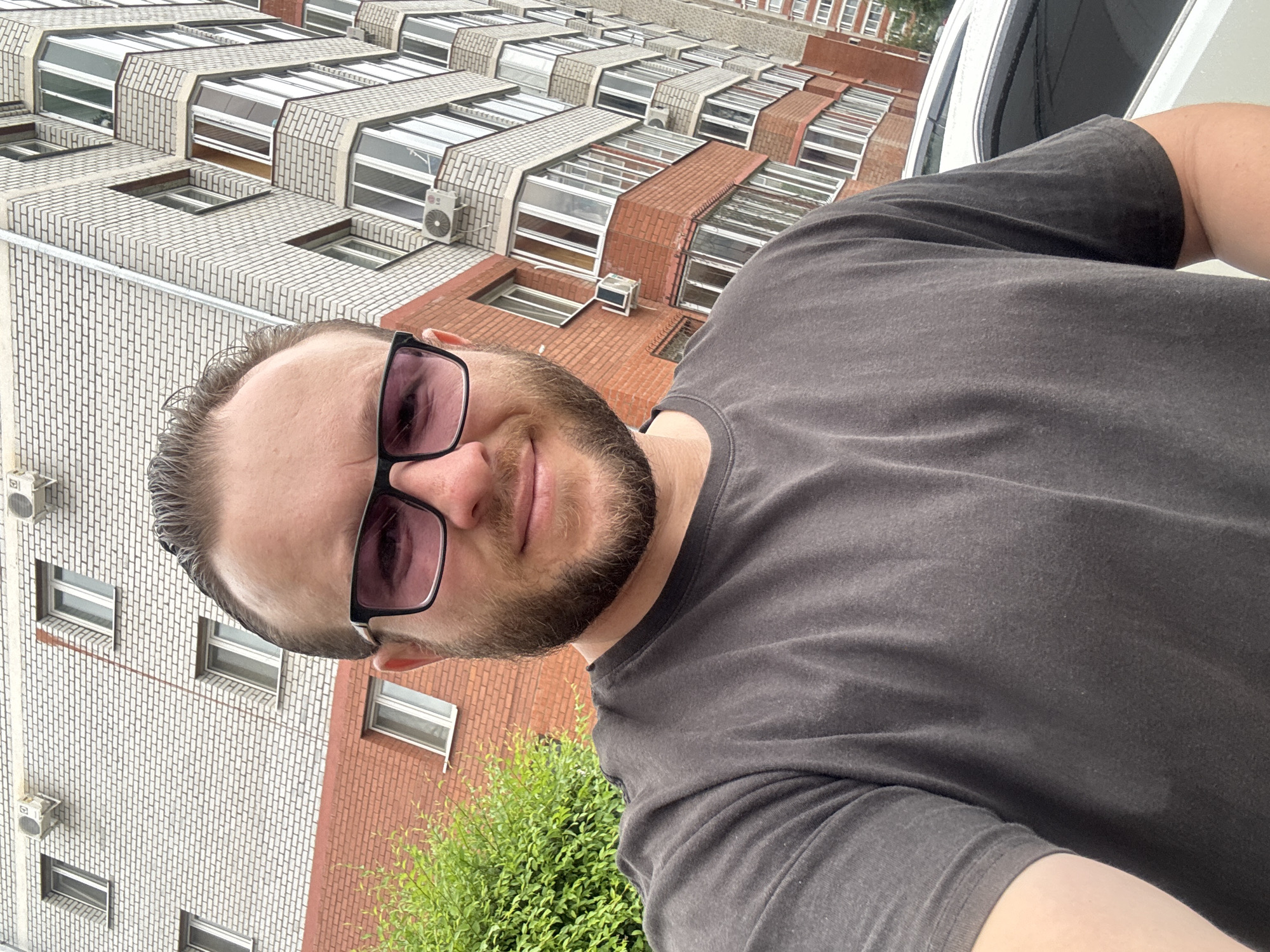 A man with a beard and short brown hair, wearing a dark gray t-shirt and dark-rimmed glasses with pink lenses, is taking a selfie outdoors. He is standing in front of a multi-story brick building, which appears to be an apartment complex. The building has numerous windows and balconies, and some air conditioning units are visible on the exterior. To the left of the man is a dense green bush. A white car is partially visible in the lower right corner of the frame. The environment suggests an urban setting. Based on visual cues such as architectural style and common building materials, the location is likely Blagoveshchensk, Russia.