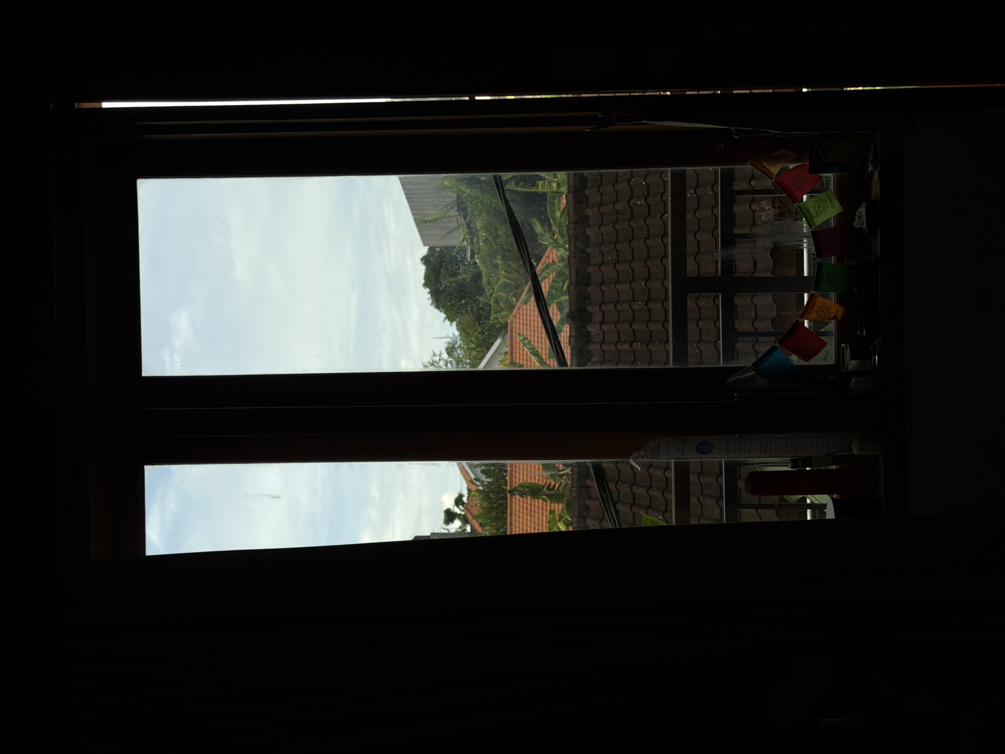 A view through a wooden-framed window from a dark room offers an outdoor scene featuring a residential area in North Kuta, Indonesia. The window, divided into two panes, reveals a partial view of the sky, appearing as a clear blue with some clouds. Beyond the window, a row of buildings can be seen. These appear to be homes with orange-tiled roofs. Behind these, there is lush vegetation, suggesting a tropical environment with trees. Colorful prayer flags are attached at the top corner of the window. A shelf is visible on the right of the window with multiple objects. The indoor space where the photo is taken is mostly dark, casting the window as a portal to the outside world.