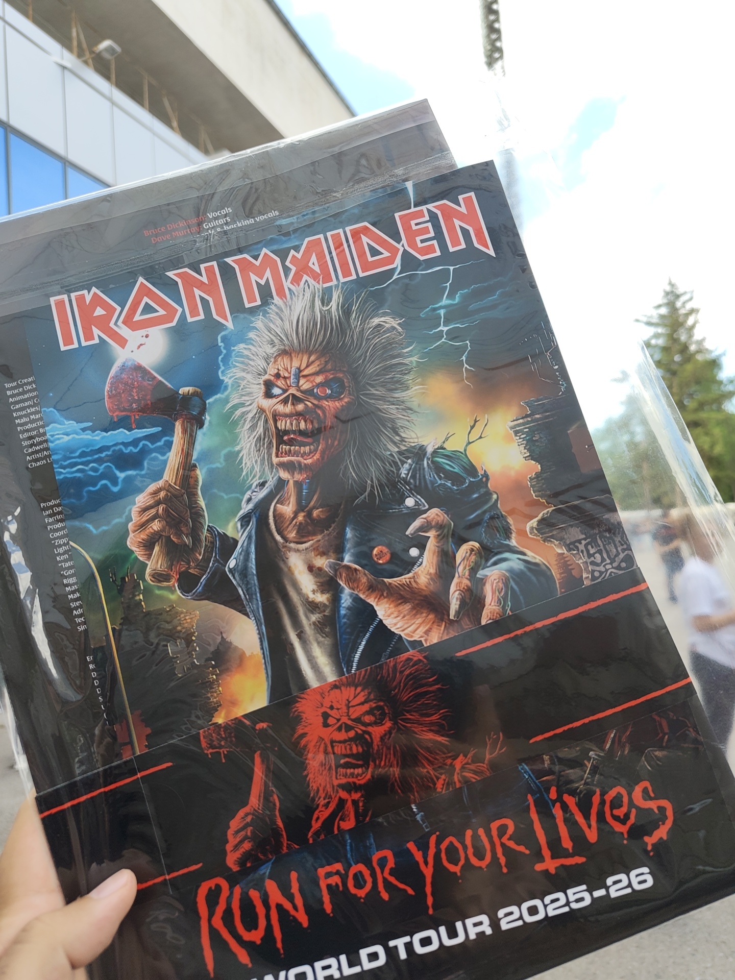 A person is holding an Iron Maiden poster titled "Run For Your Lives WORLD TOUR 2025-26" in front of a modern building with glass windows. The poster features the band's mascot, Eddie, in two different guises. A person in a white shirt is walking in the background. There are trees and a light pole in the background. The poster lists Bruce Dickinson as vocals and Dave Murray as guitars. The building appears to be in an urban setting, potentially in Sofia, Bulgaria, though the specific location isn't discernible from the image.
