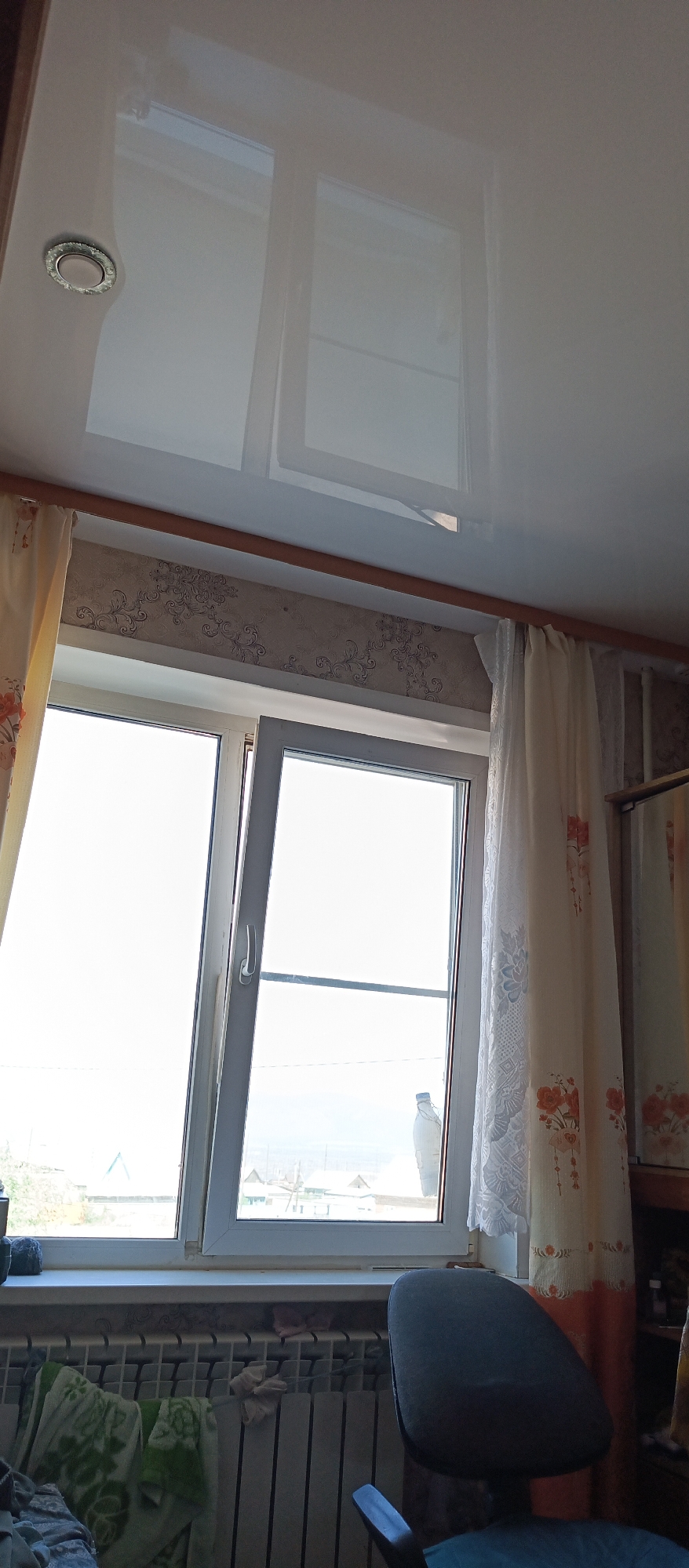 A view from inside a room in Gusinoozyorsk, Russia, looking towards a window. The room has a white, glossy, stretch ceiling with integrated lighting. A wooden curtain rod runs across the top of the window. On the left, a sheer yellow curtain with an orange floral pattern hangs. On the right, a white sheer curtain with an intricate lace pattern and a similar orange floral trim is visible. The window itself is a modern white plastic frame with two panes. The left pane is open outwards, showing a bright, overcast sky and a distant landscape with some buildings and trees. The right pane is closed, and a blue bottle is visible on the windowsill outside. Below the window is a white windowsill and a white radiator. A dark grey office chair is positioned in the foreground, partially obscuring the lower right portion of the radiator. There are some items on the radiator, including a green and white patterned fabric and a light-colored cloth. The wallpaper above the window has a subtle pattern of swirling grey designs. The overall impression is an indoor domestic scene on a cool, possibly winter day, given the presence of a radiator and the overcast sky.