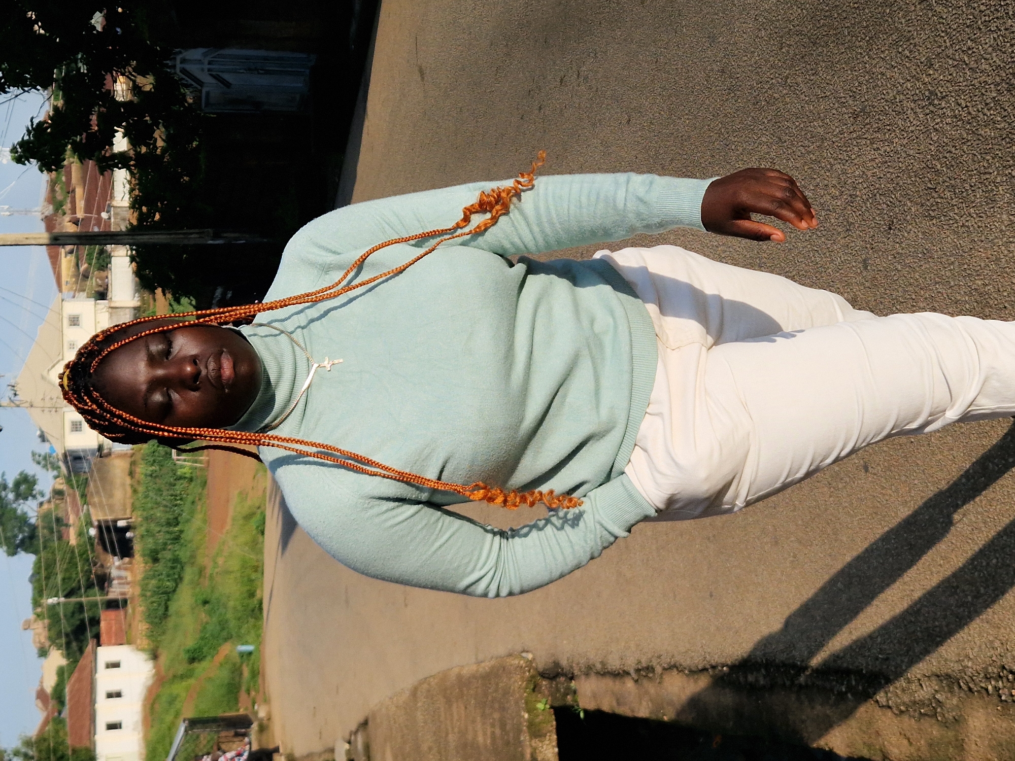 A Black woman with braided hair walks down a paved road in Kabong, Nigeria. She is wearing a light blue turtleneck sweater and cream-colored pants. The woman has her eyes closed and appears to be looking down. A thin silver chain with a cross pendant is visible around her neck. The road is lined with a concrete curb on the left side and vegetation and buildings on the right. The sun casts shadows on the road. In the background, there are houses, trees, and utility poles. The sky is clear and blue.