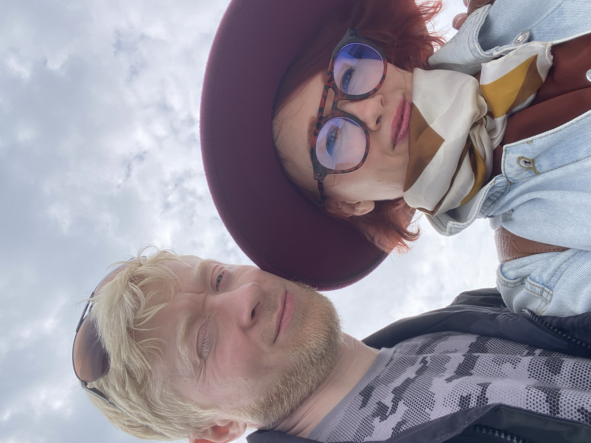 From a low angle, a fair-skinned man and a fair-skinned woman are facing the camera. The man has blond hair, a beard, and sunglasses on his head. He is wearing a gray patterned shirt with a black jacket over it. The woman has red hair, wears a dark red hat, and glasses. She has on a blue denim jacket over a brown shirt and a patterned scarf. The background is a cloudy sky. No other clues suggest that this photo was taken in Sochi, Russia.