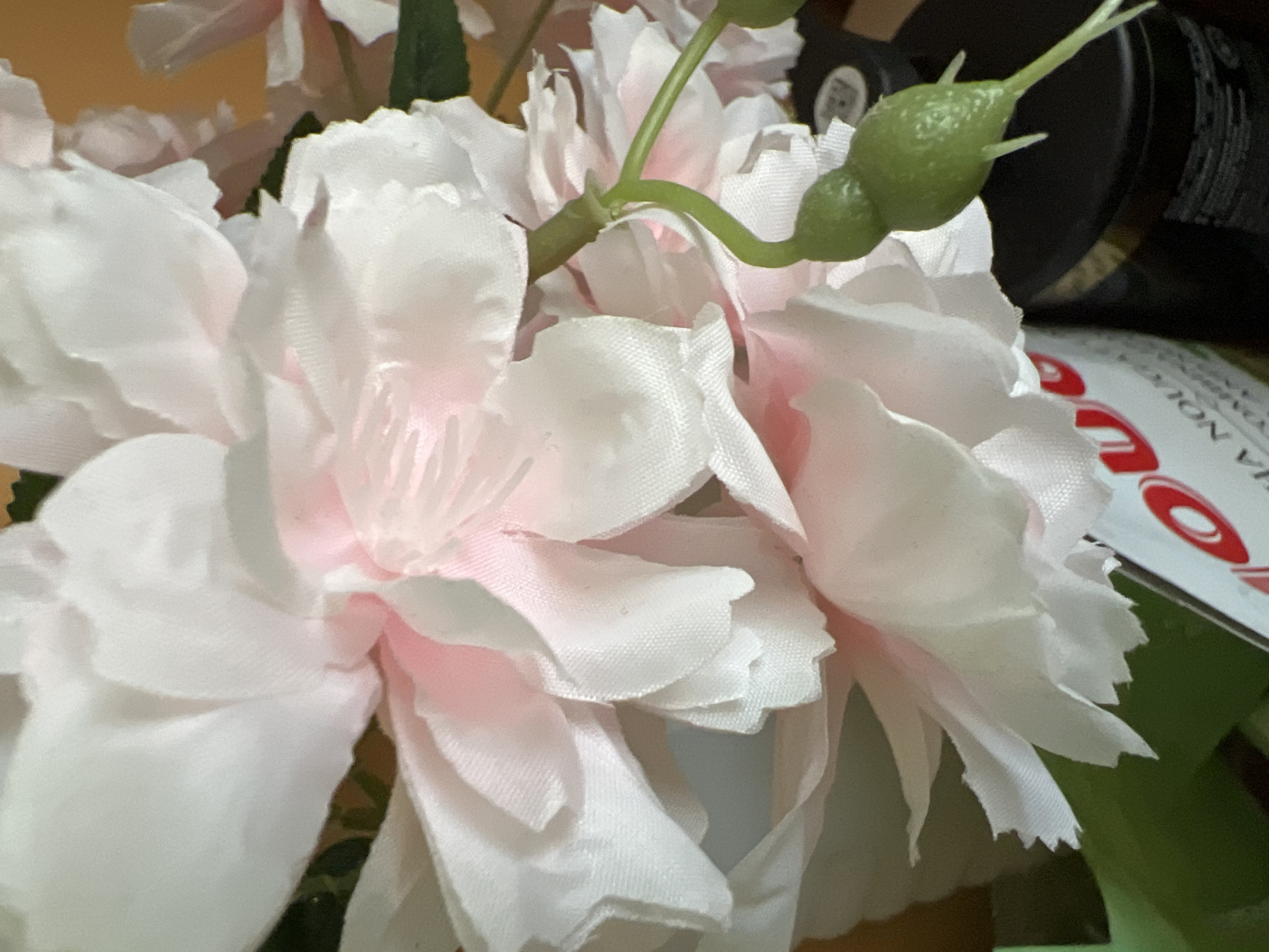 The image shows a close-up of artificial white and pink flowers with a green stem and two small green buds. In the background, there are blurred objects, including a dark cylindrical container and part of a white label with red lettering, possibly from a product. The lighting suggests an indoor setting. There are no people or animals visible in the image. The image does not contain any visual information that would allow for its location to be identified as Minsk, Belarus.