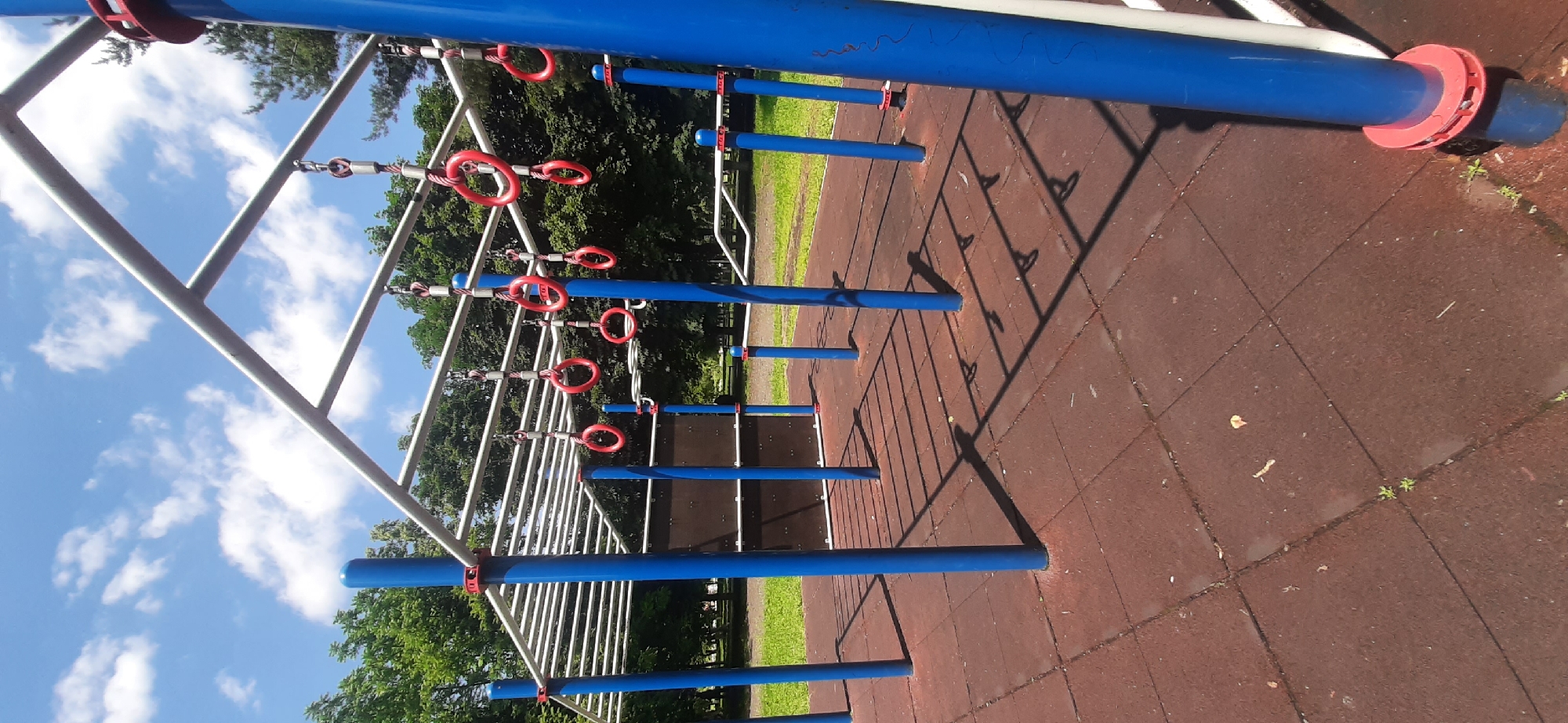 An outdoor exercise park is depicted under a partly cloudy sky in Yessentukskaya, Russia. The foreground consists of a red rubberized mat surface with a tiled pattern. The park features a metal structure with blue support poles and horizontal bars. Several red rings are suspended from chains attached to the horizontal bars, suggesting equipment for gymnastics or calisthenics. In the background, lush green trees and a line of deciduous trees form a natural backdrop. The lighting suggests it is daytime, with clear shadows cast by the structures. There are no people or animals visible in the scene.