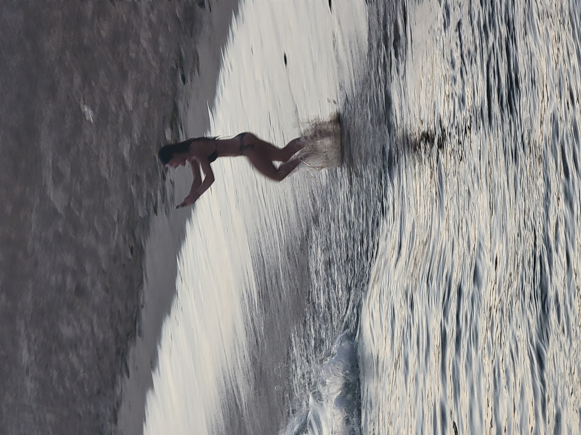 A woman in a bikini is captured mid-stride on a sandy beach in Quarteira, Portugal. She is holding a smartphone in her right hand, appearing to record or take a photo, with her mouth open in an exclamation. Water splashes around her feet as she moves from the sea onto the shore. The background consists of the grey, textured sand of the beach, transitioning into the darker, rippling surface of the ocean in the foreground. The lighting suggests it is either early morning or late evening, casting long shadows and creating a muted color palette.