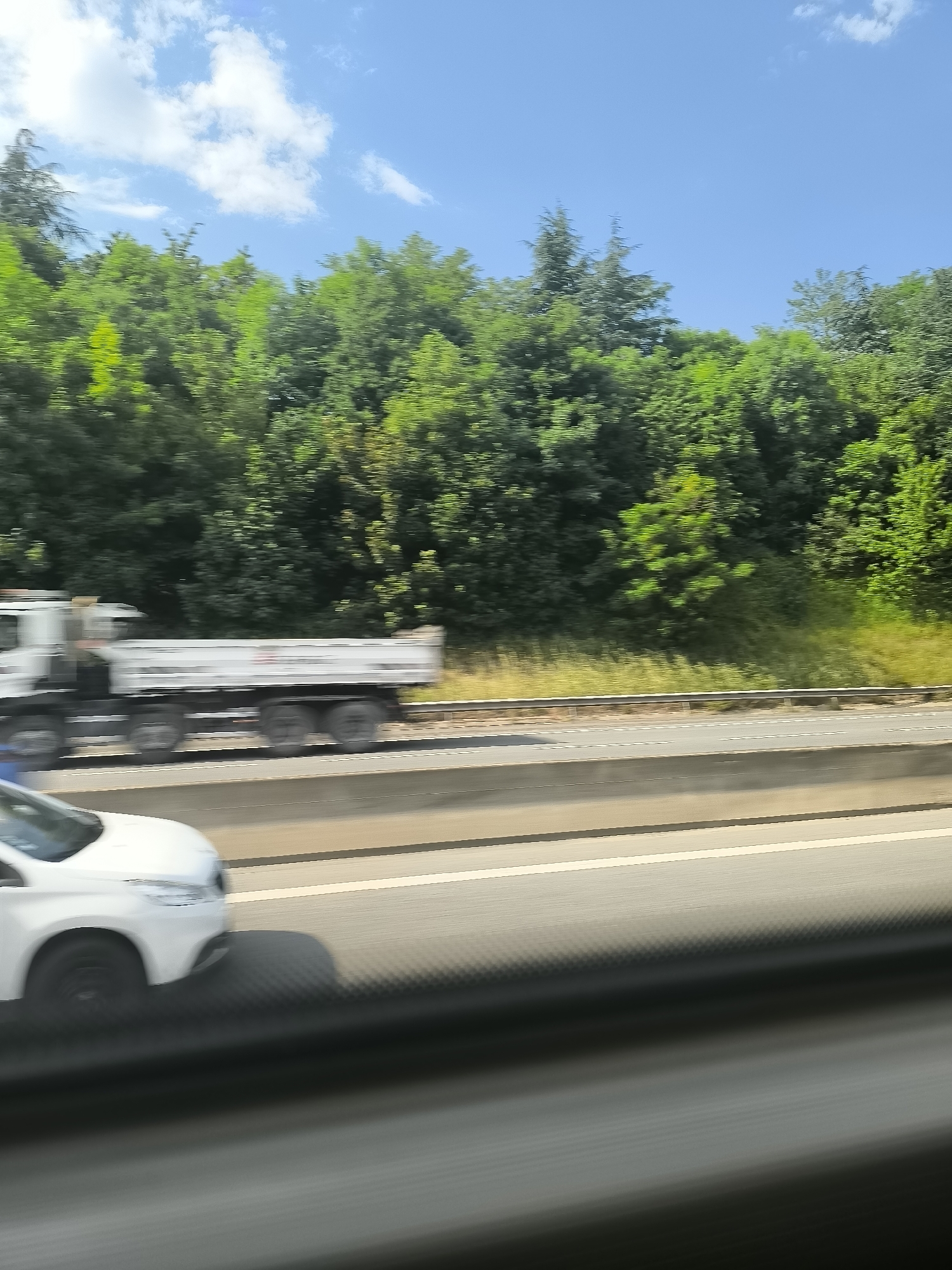The image shows a blurry view from a moving vehicle on a highway. In the foreground, part of a white car is visible, moving in the same direction as the vehicle from which the picture is taken. On the left side of the highway, a large white truck with a flatbed is speeding by. Beyond the highway, a dense line of green trees extends across the horizon under a blue sky with scattered white clouds. The location can be deduced to be near Saint-Jean-Bonnefonds, France based on similar landscape and architectural factors, which indicates that the picture was taken in a rural or suburban environment with close highway access.