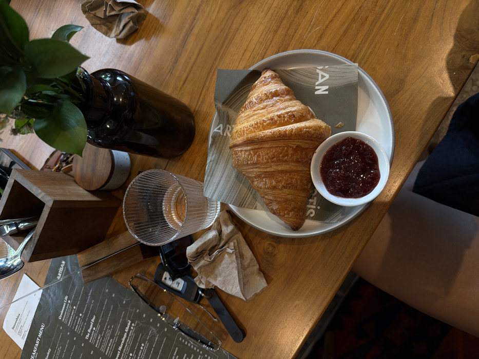 A top-down shot reveals a rustic wooden table setting. A croissant, accompanied by a small bowl of jam, rests on a plate with branding. Nearby, a glass cup and a dark brown bottle with greenery add to the scene. A crumpled napkin and a menu with cutlery lie on the table, completing the breakfast arrangement. A portion of a person's clothing is visible in the corner.
