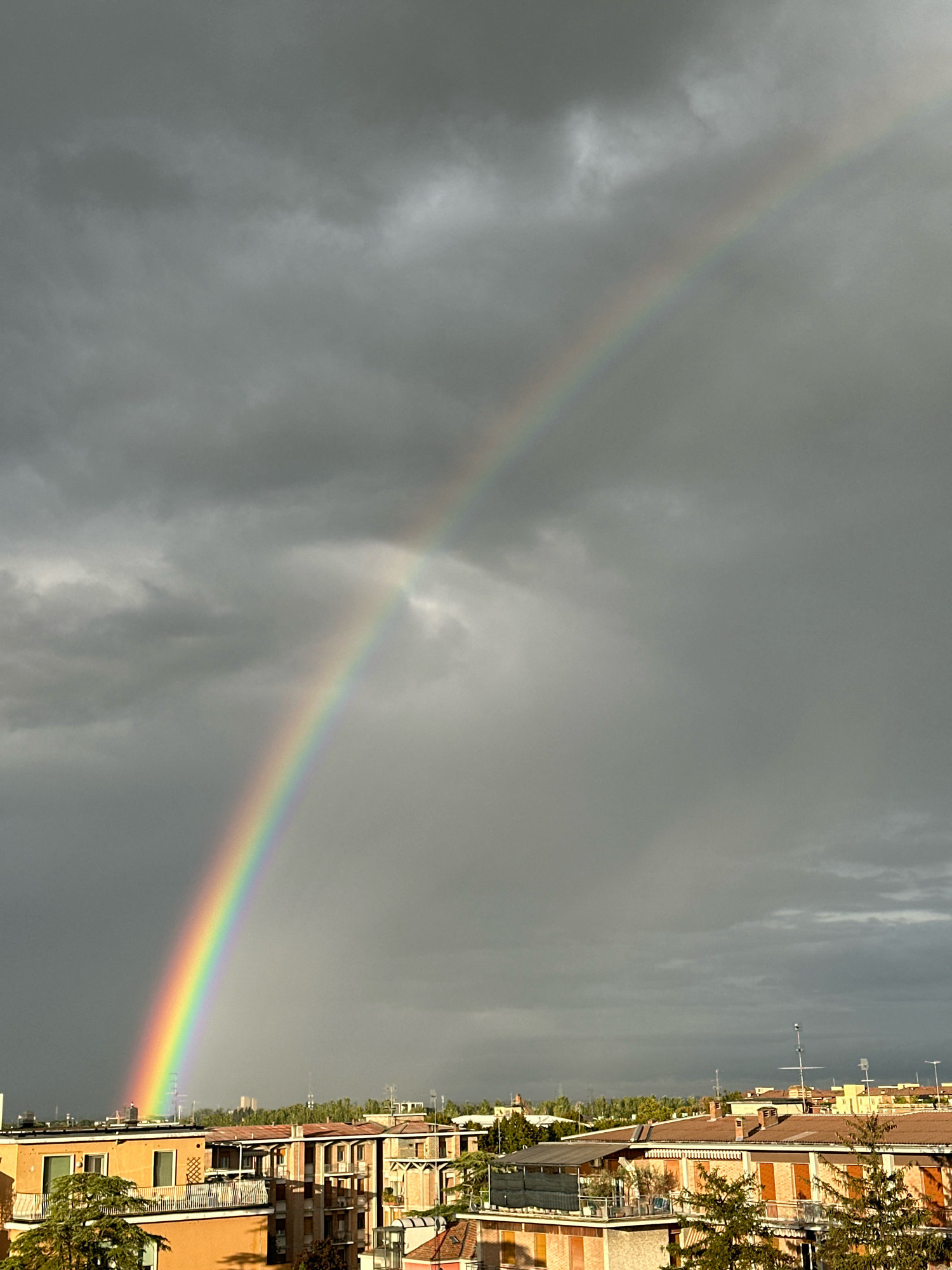 A wide shot captures a vibrant rainbow arcing across a dramatic, overcast sky over the city of Parma, Italy. The rainbow, a spectrum of vivid colors, originates from the left side of the frame and ascends towards the upper right. Below the sky, the rooftops of apartment buildings are visible, characterized by their terracotta tiles and various architectural styles. Green trees dot the landscape between the buildings. The scene suggests a recent rain shower, with the dark clouds and the subsequent appearance of the rainbow indicating a shift in weather. No people or specific actions are discernible in the image.