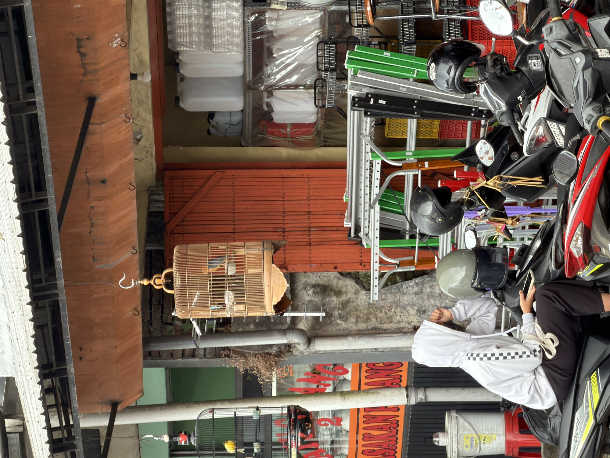 In the city of Kintamani, Indonesia, a person wearing a white hooded sweatshirt and black pants sits on a black motorcycle. The person is holding a mobile phone and appears to be looking away from the camera. The motorcycle is parked in front of a store with an orange corrugated metal door. Two birdcages hang from an awning above the store. One cage contains a yellow bird, and the other contains two birds, one of which appears to be blue and orange. Various items are displayed on shelves behind the open door, including white plastic containers, red and white bags in plastic wrapping, and what appear to be stacked plastic crates in yellow and red. Several other motorcycles are parked nearby, some with helmets resting on them. The ground is paved, and the overall setting suggests an outdoor market or commercial area.