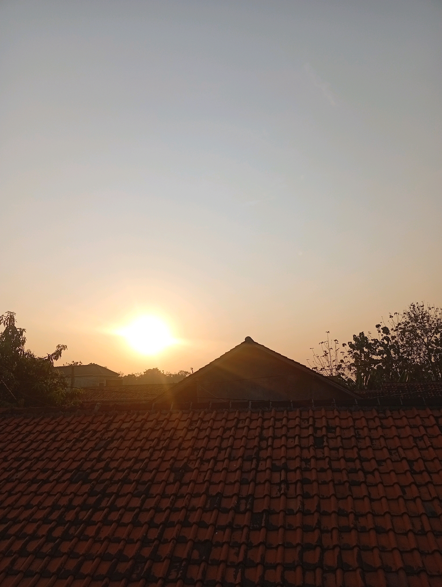 The image depicts a rooftop view of houses in Kebandingan, Indonesia, during either sunrise or sunset. The foreground is dominated by a weathered terracotta tiled roof, showing signs of age and some discoloration. Beyond the foreground roof, a darker, pitched roof of a building is visible, with the setting or rising sun casting a warm, golden glow behind it. Silhouetted trees and other foliage are scattered across the middle ground, interspersed with the roofs of other houses. The sky above is a gradient of pale blue transitioning to a warm orange hue near the horizon, where the sun is prominently positioned. The overall impression is one of a serene, residential scene at dusk or dawn.
